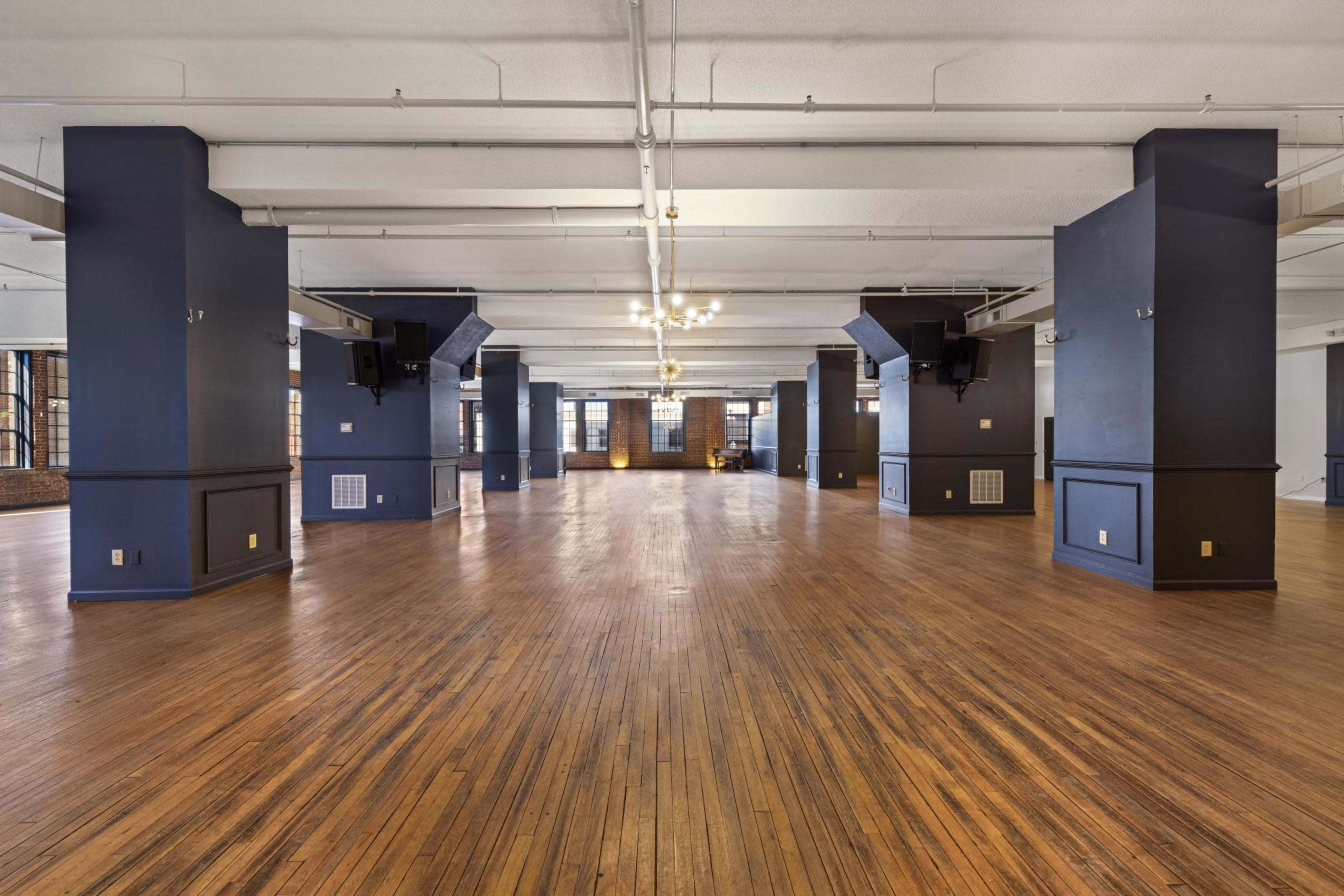 A spacious, empty room with wooden flooring and large windows, featuring dark-painted columns.