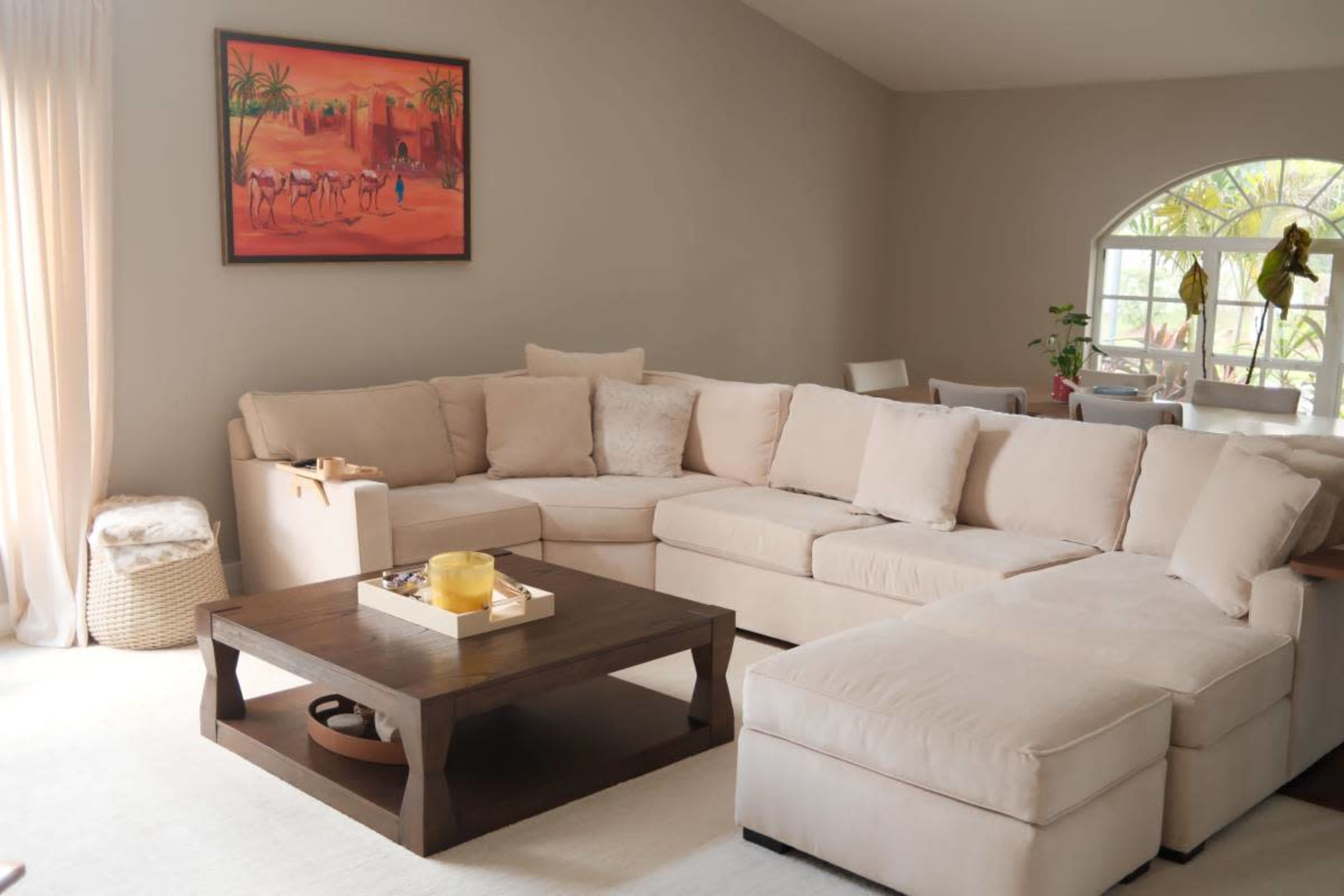 The image shows a light-colored, spacious living room with a large sectional sofa, a wooden coffee table, and a wall-mounted painting depicting a desert scene.