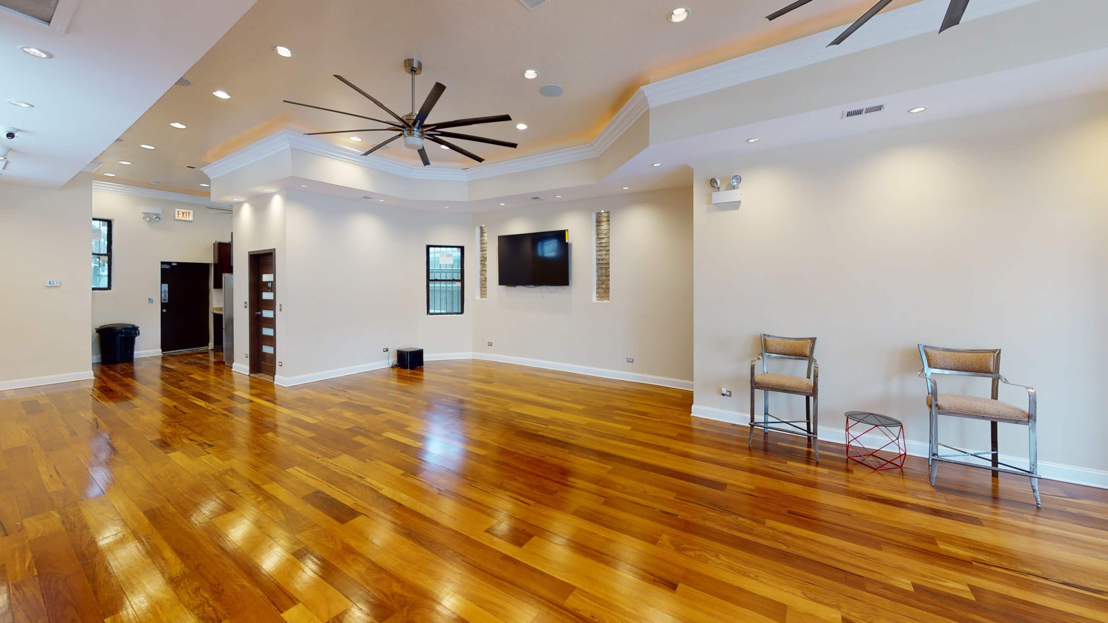 A spacious room with polished wooden floors, ceiling fans, a mounted television, and two chairs positioned near a side table.