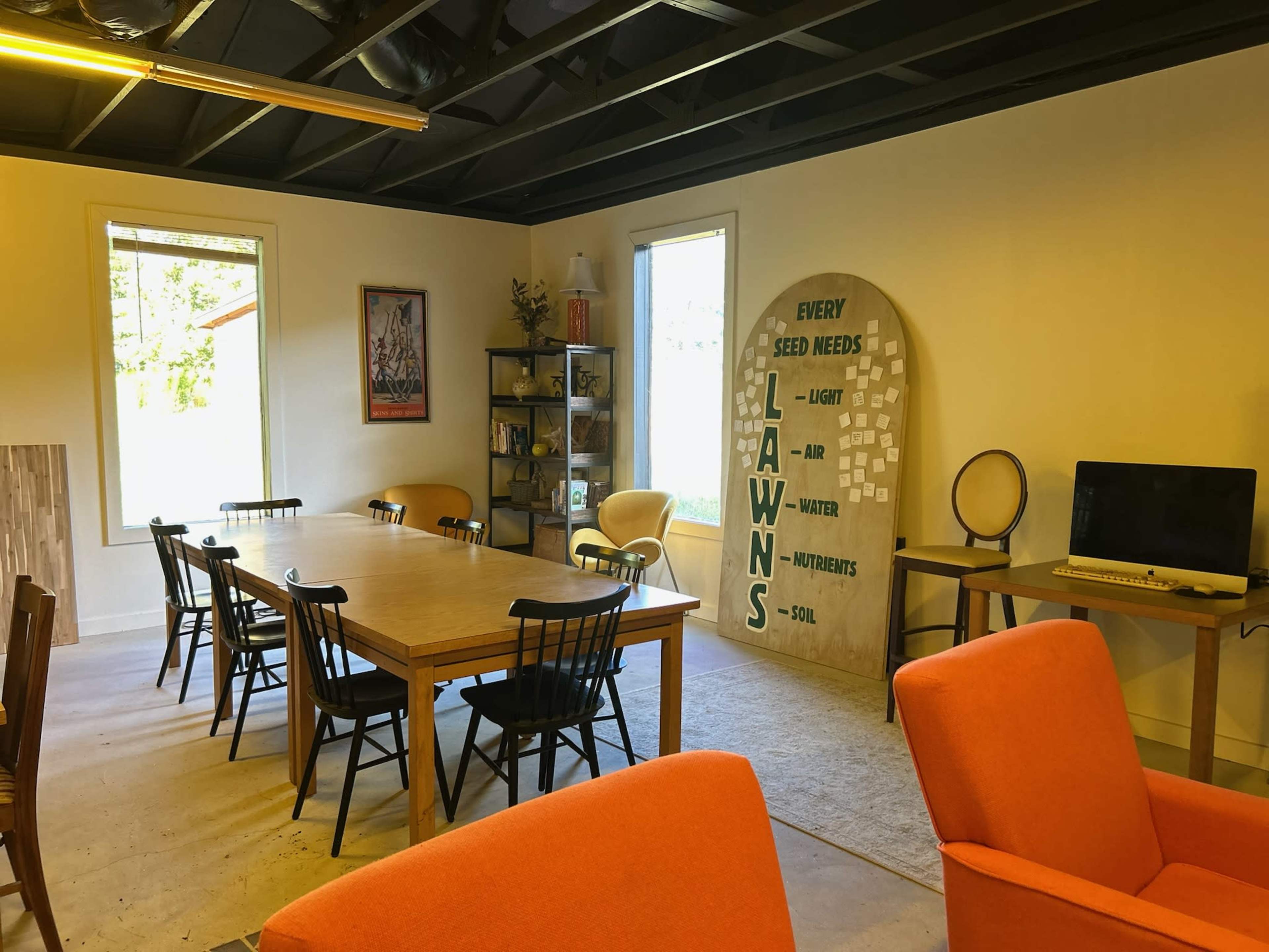 The image shows a spacious room with a large wooden table, black chairs, orange armchairs, and a wall featuring the phrase "Every Seed Needs" next to a list of essential elements for growth.