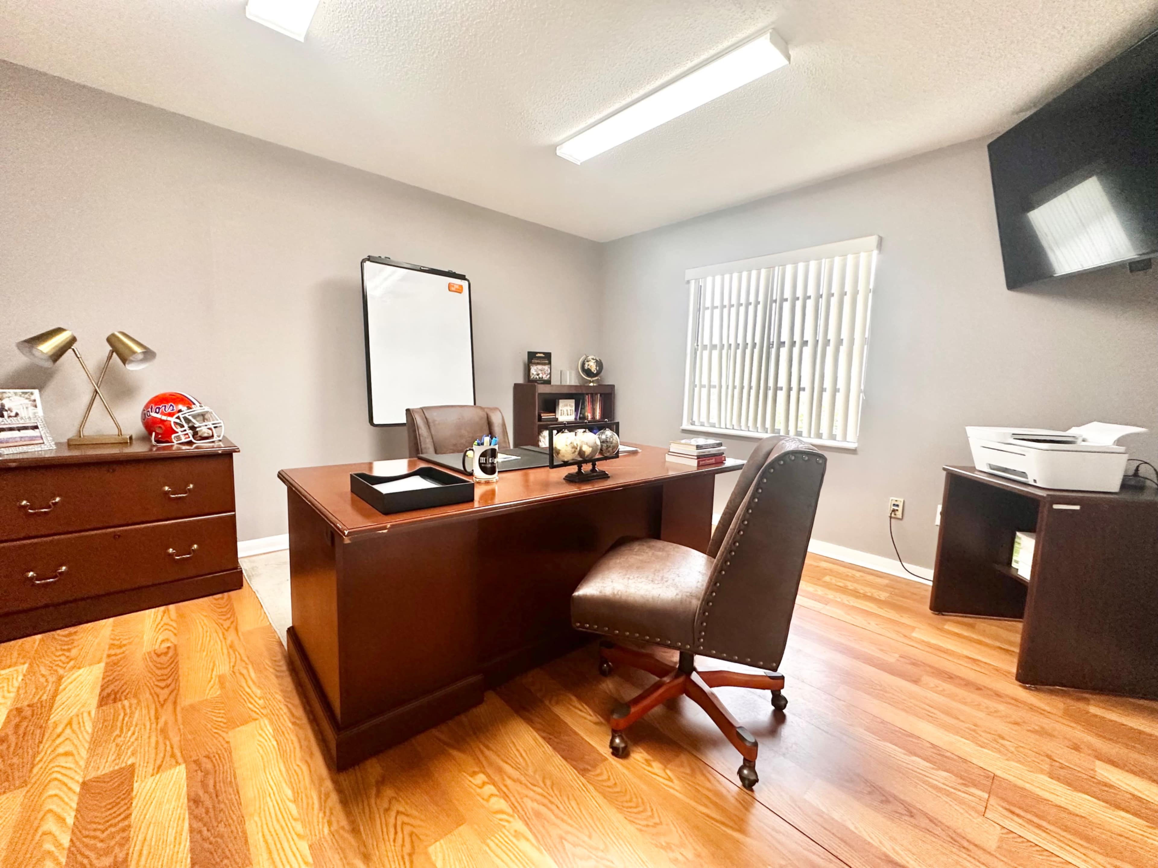 The image shows a modern office with a brown wooden desk, an ergonomic chair, a whiteboard, a printer, and framed pictures on the walls.