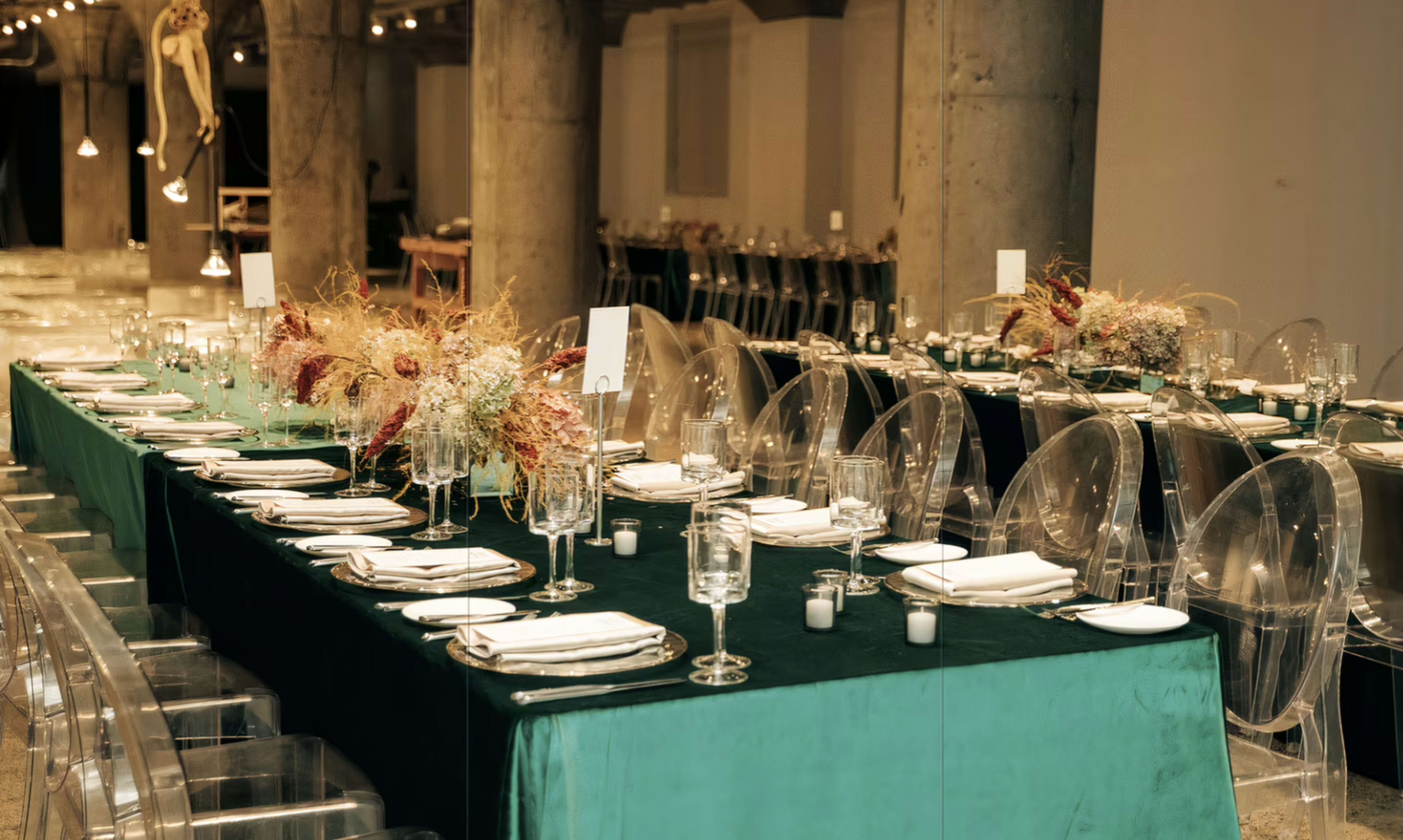 A long dining table is set with glassware and silverware, draped in a green tablecloth and adorned with floral arrangements, in a spacious indoor venue.