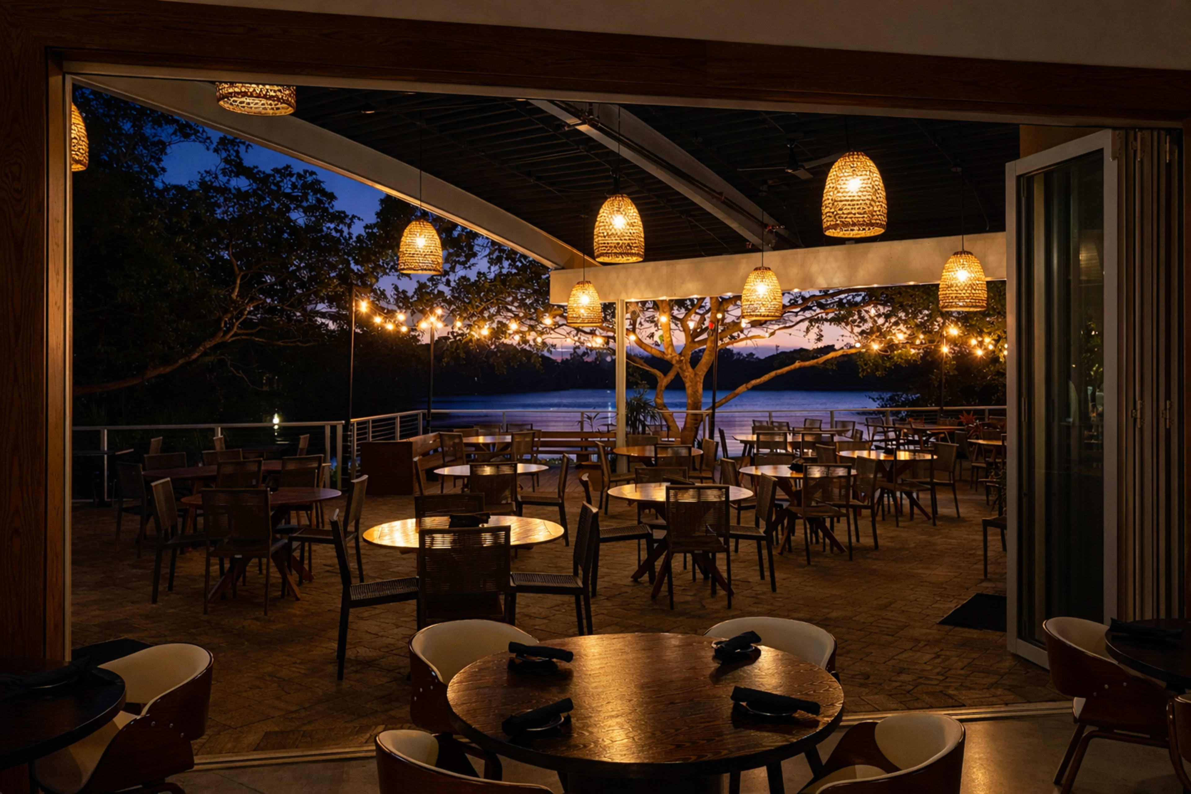 The image shows a partially enclosed restaurant space featuring empty tables and chairs with warm pendant lighting, overlooking a tranquil waterfront at dusk.