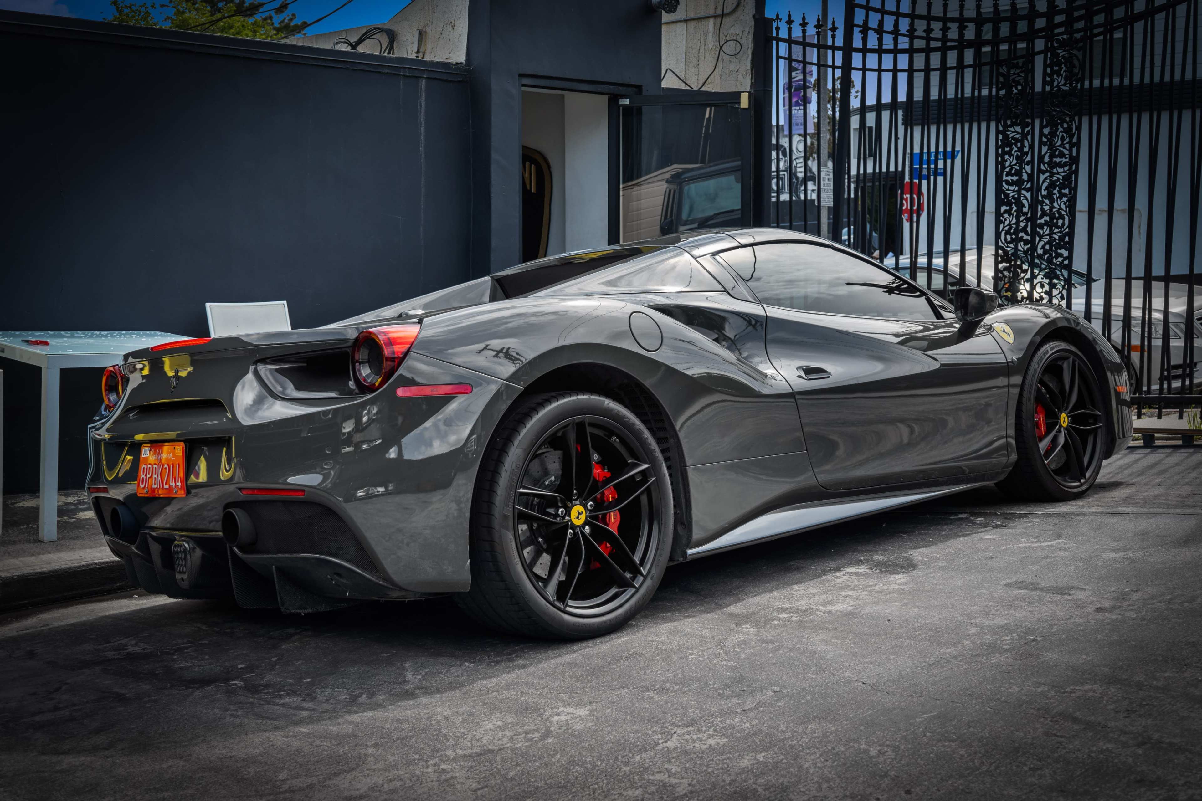 A gray Ferrari 488 GTB is parked on a street beside a black gate.