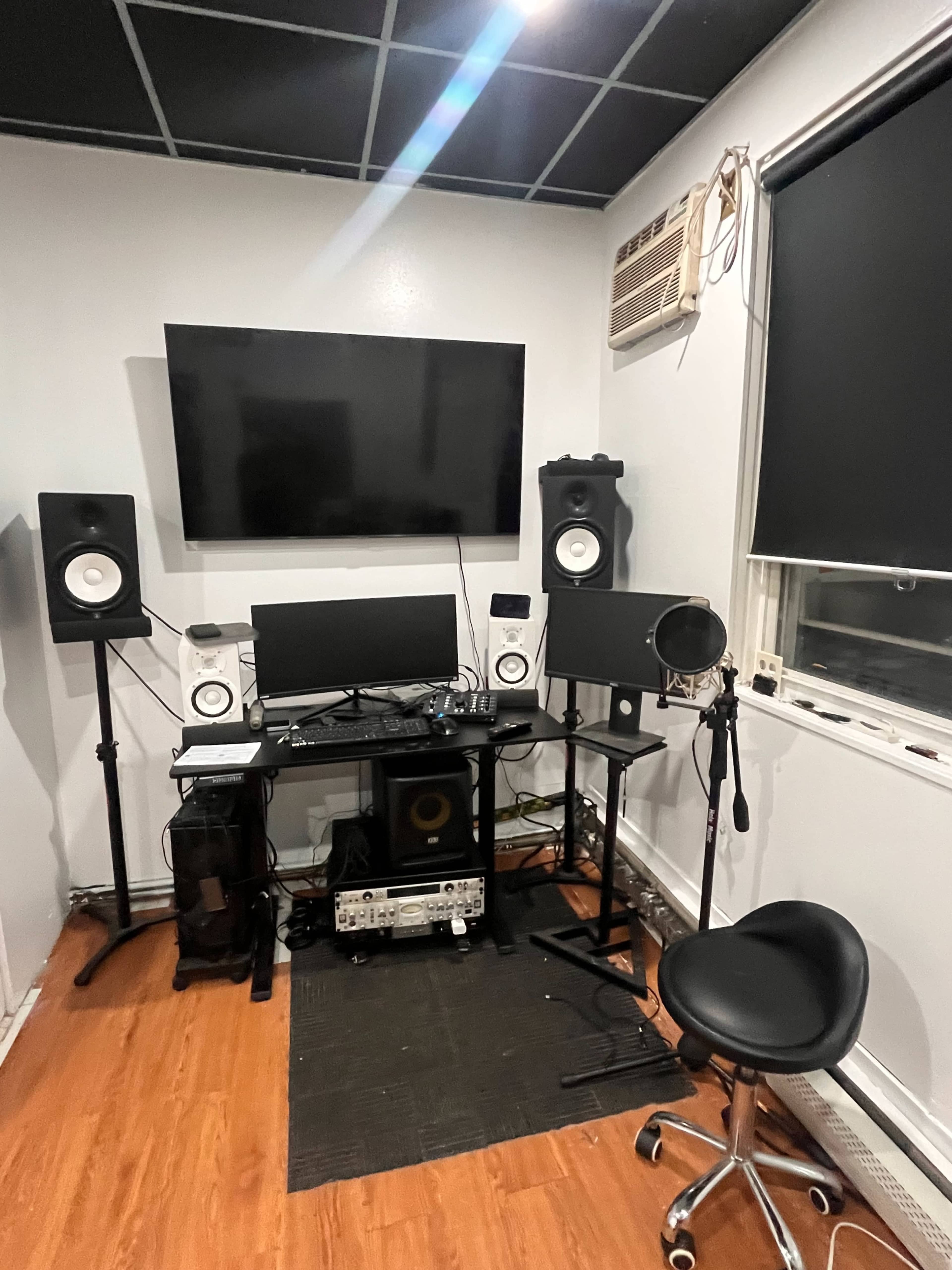 The image shows a small music studio setup featuring a desk with monitors, speakers, a microphone on a stand, and a large television mounted on the wall.