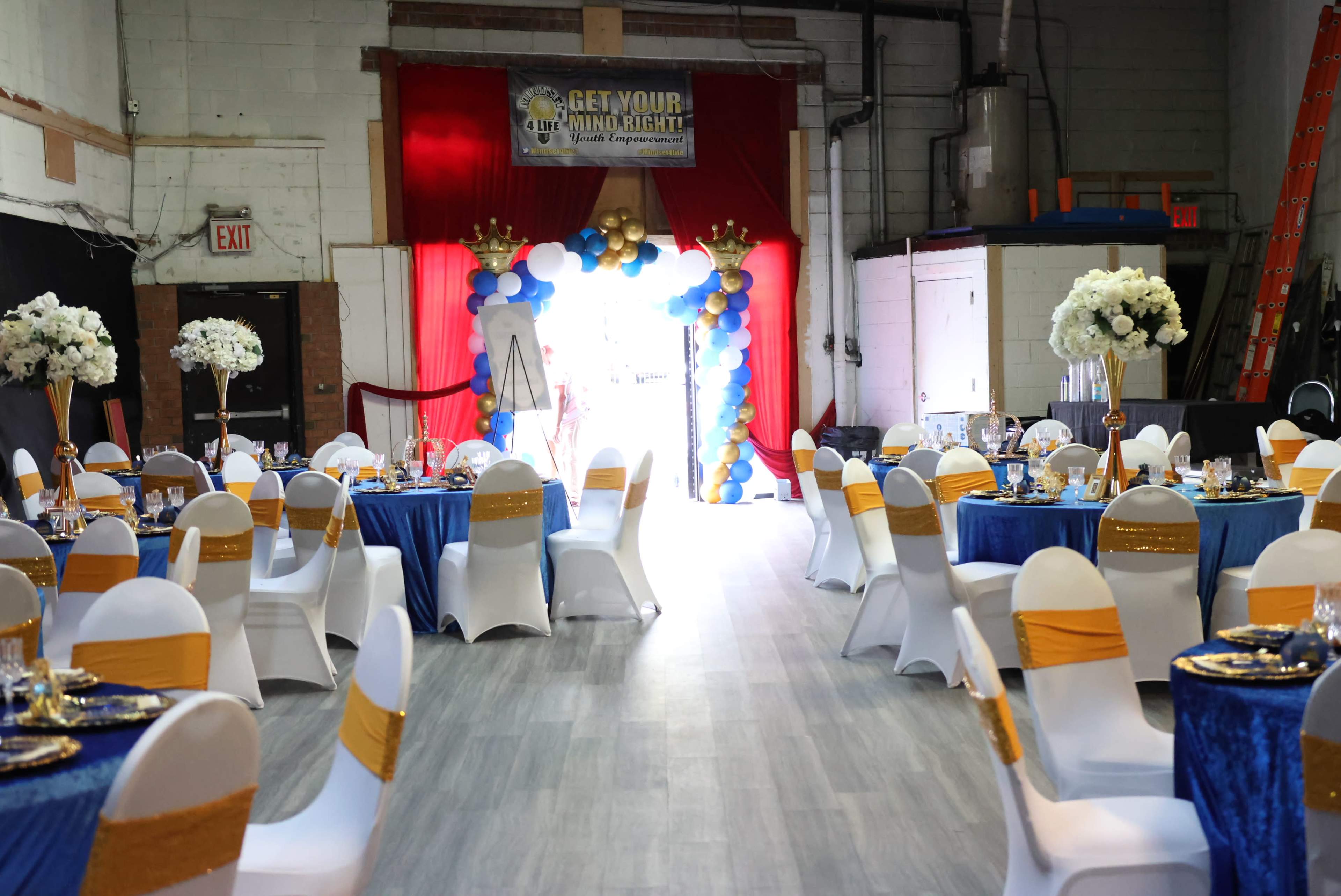 The image shows an event venue arranged with round tables covered in blue and gold tablecloths, surrounded by white chairs decorated with gold sashes, and a decorated entrance featuring blue and gold balloons.