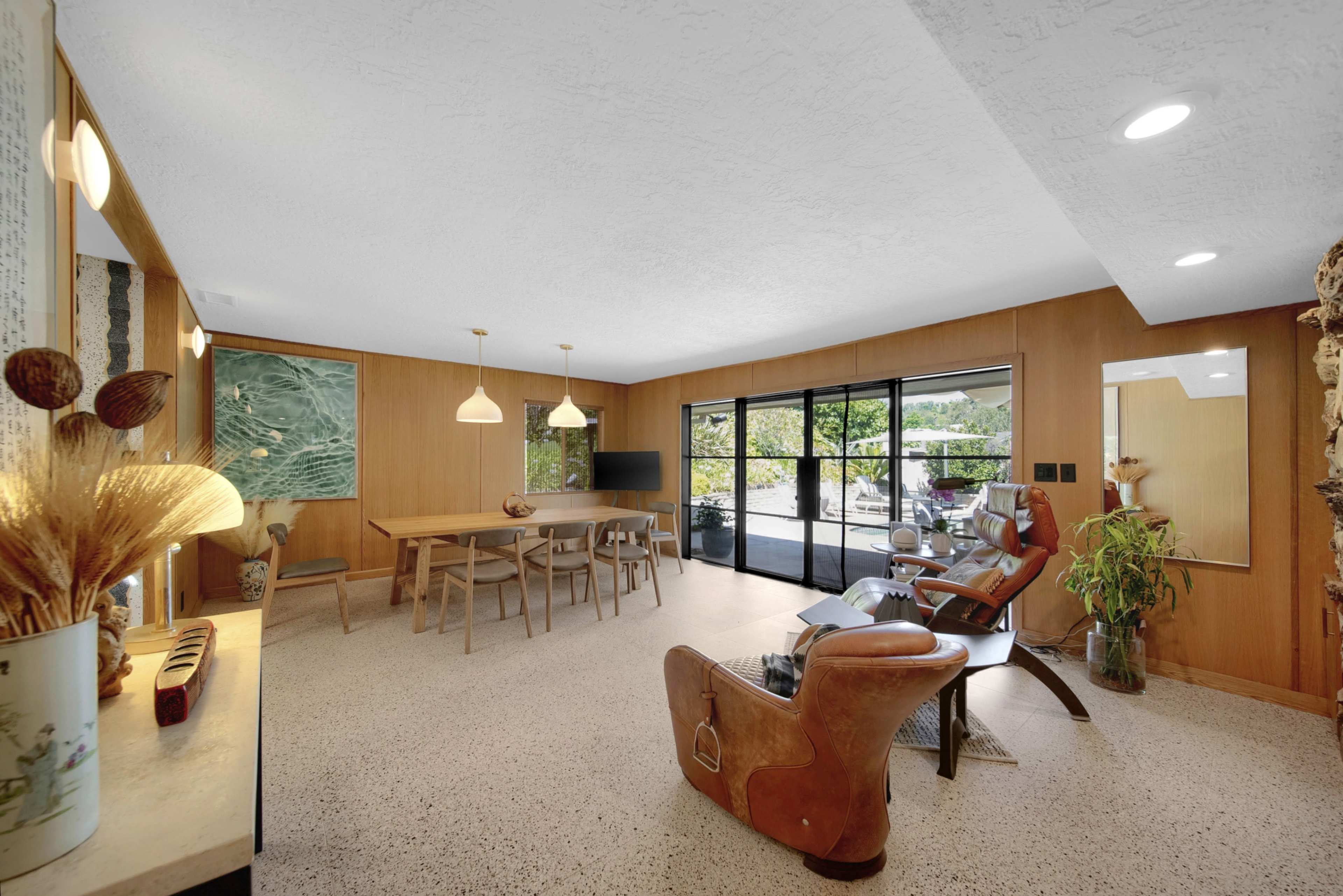 The image shows a modern living space with wooden walls, a dining area with a table and chairs, a seating area featuring a reclining chair, and large glass doors leading to an outdoor patio.