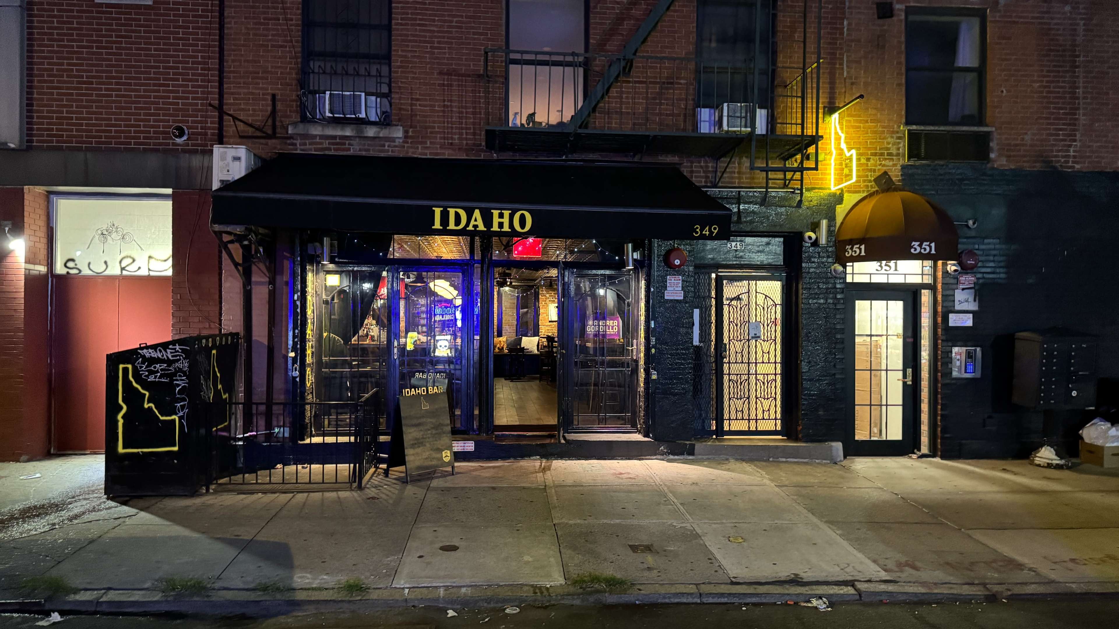 The image shows a bar named "Idaho" with a black awning, located on a street marked by a visible dumpster and neighboring buildings, at night.