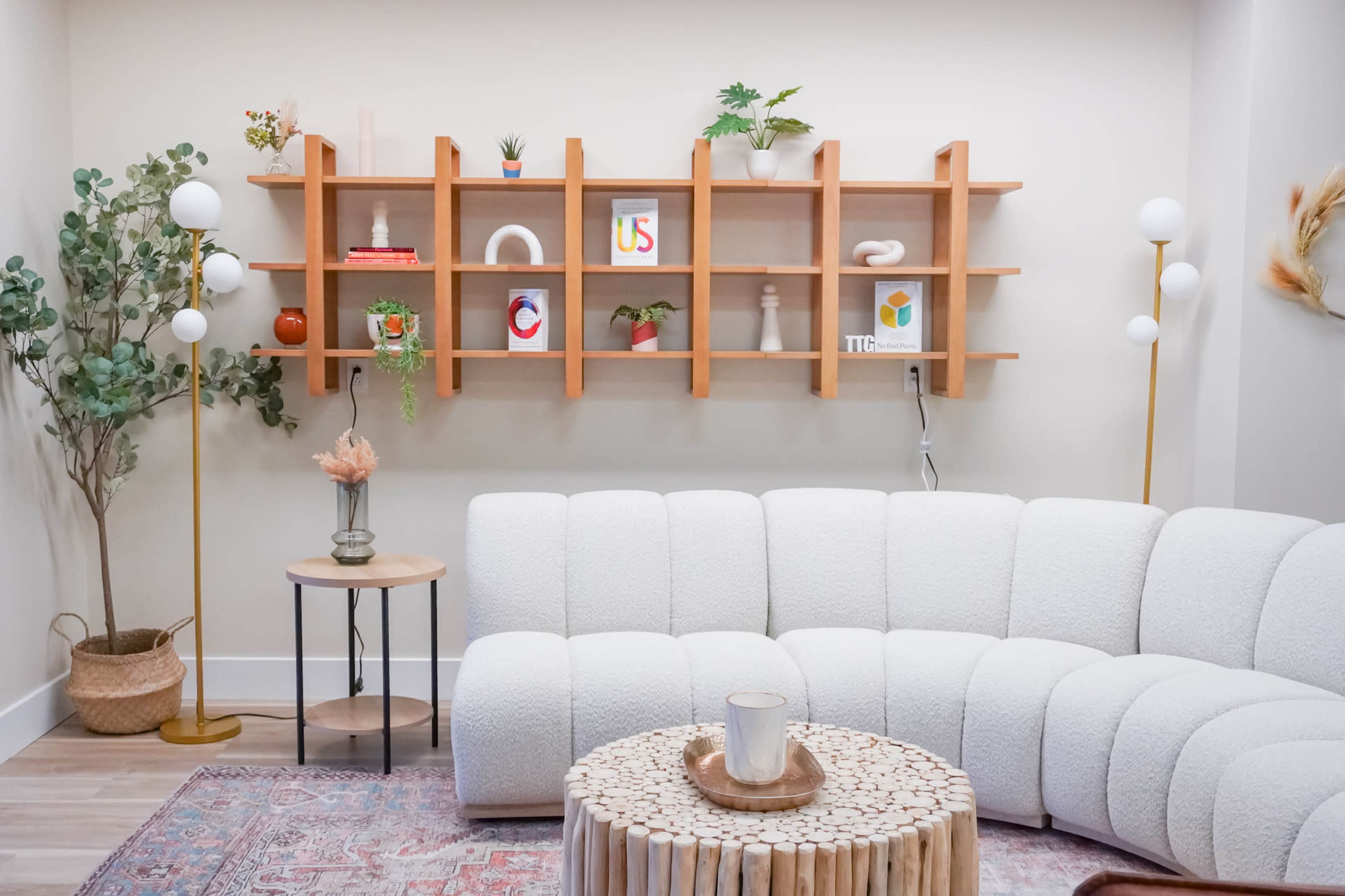 The image shows a cozy living room with a curved white couch, a decorative wooden shelf filled with plants and objects, and a round coffee table made of stacked wooden pieces.