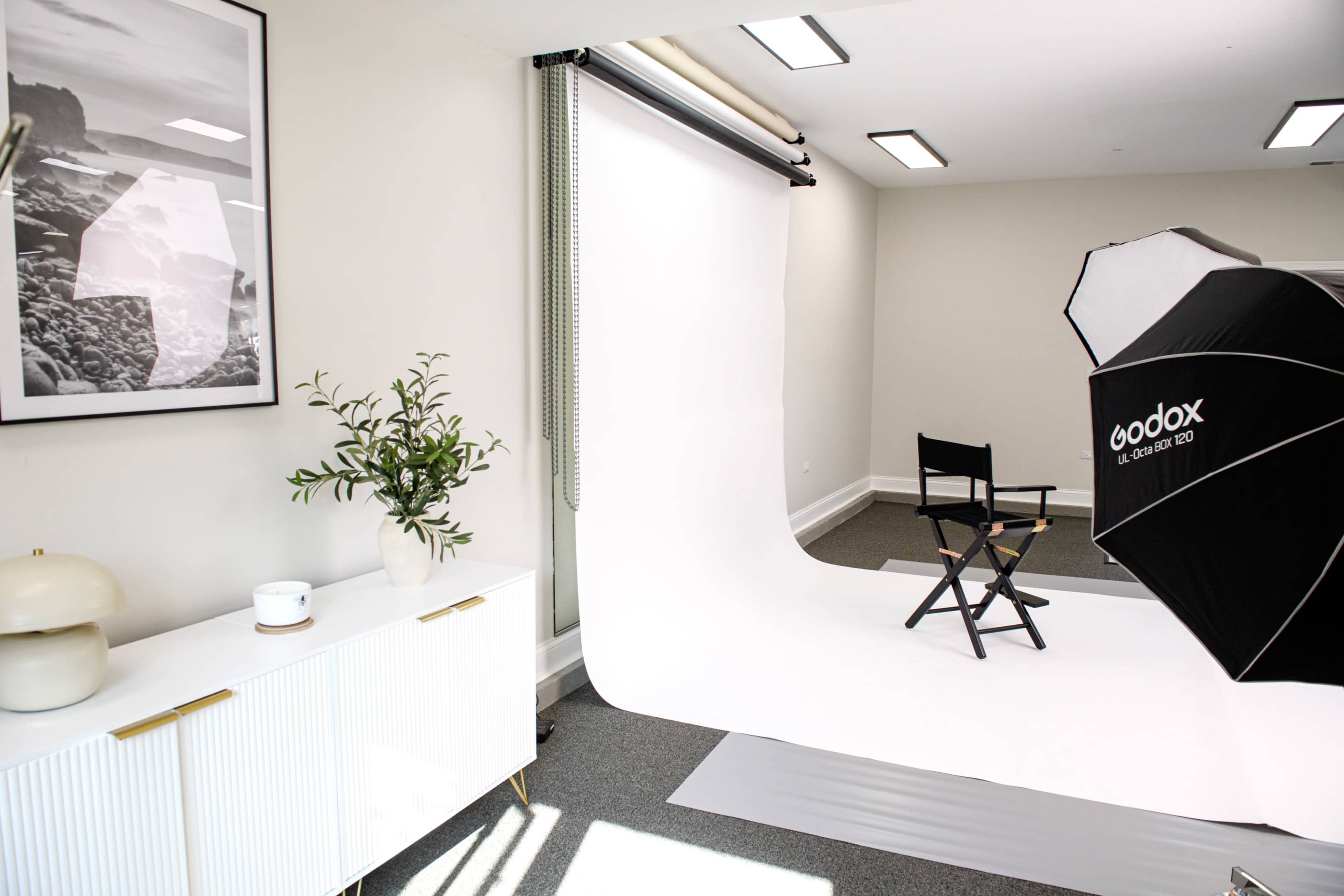 The image shows a bright studio space with a white backdrop, a director's chair, and lighting equipment, alongside a modern sideboard and a framed landscape photograph on the wall.