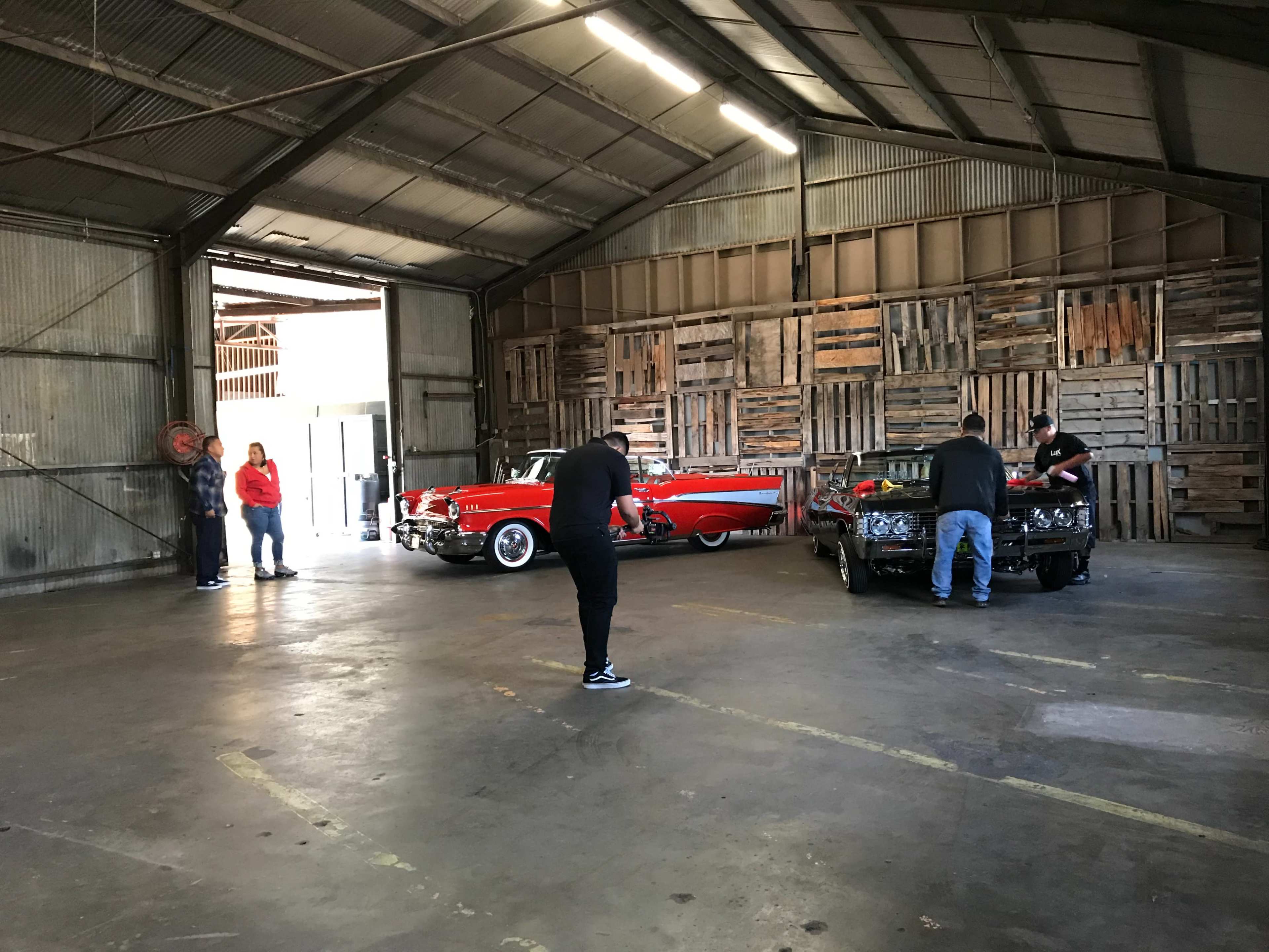 The image shows a spacious warehouse with two classic cars, a red convertible and a black vehicle, while several people are engaged in various activities around them.