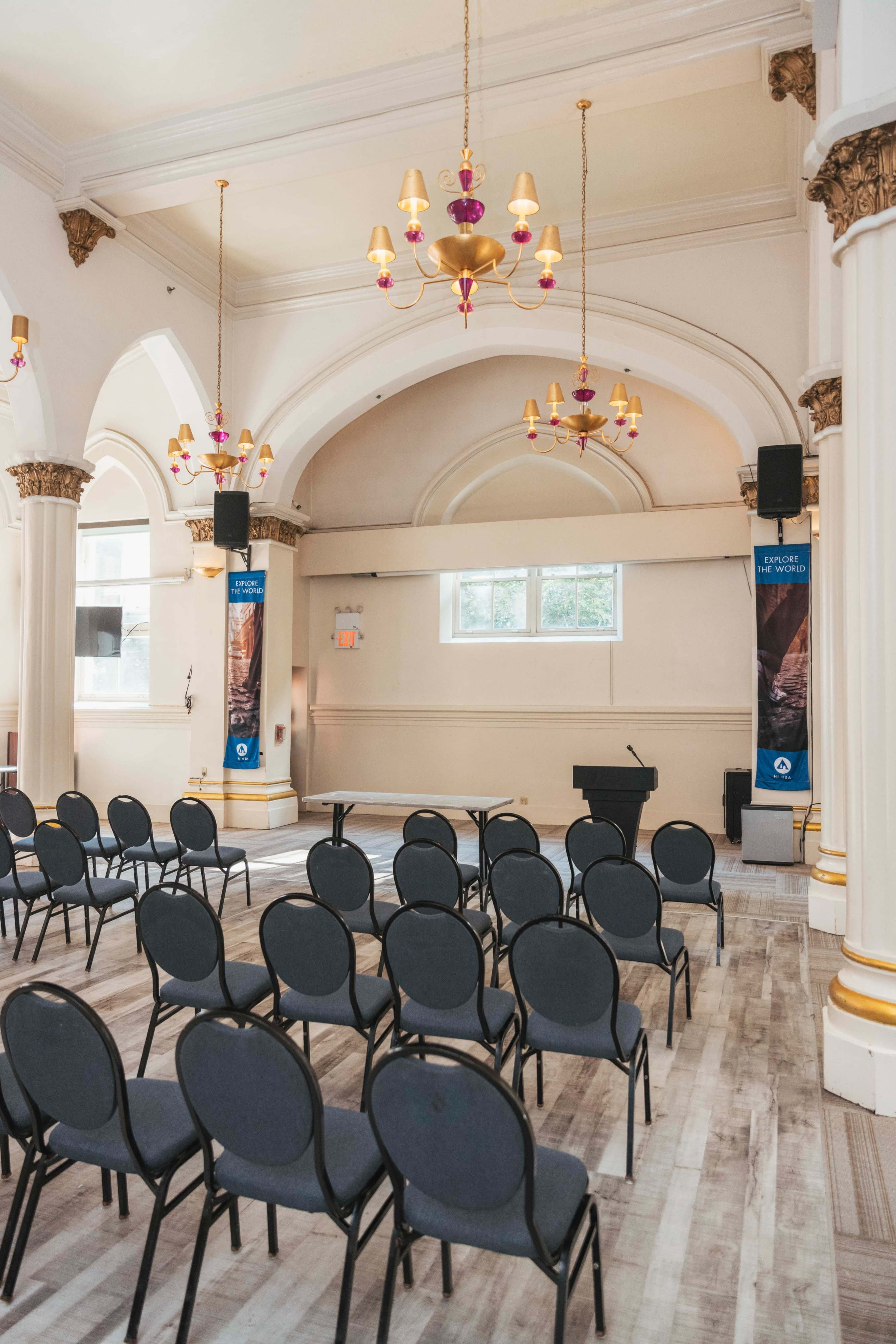 The image shows a spacious room set up for an event, featuring rows of chairs facing a podium, with decorative chandeliers and arched windows.