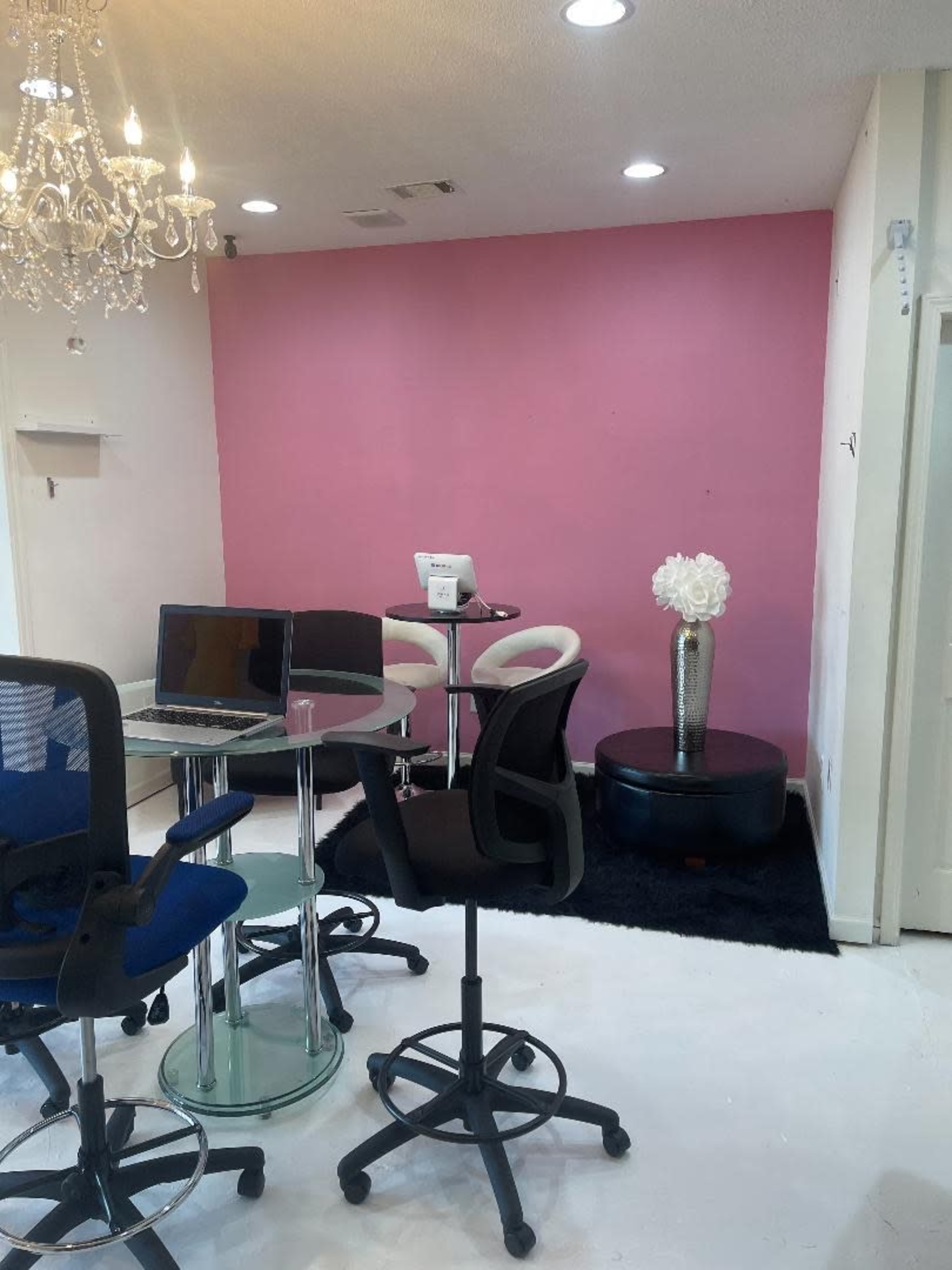 A modern office space featuring a pink accent wall, a glass table with chairs, a black ottoman, and a decorative vase with a white flower.
