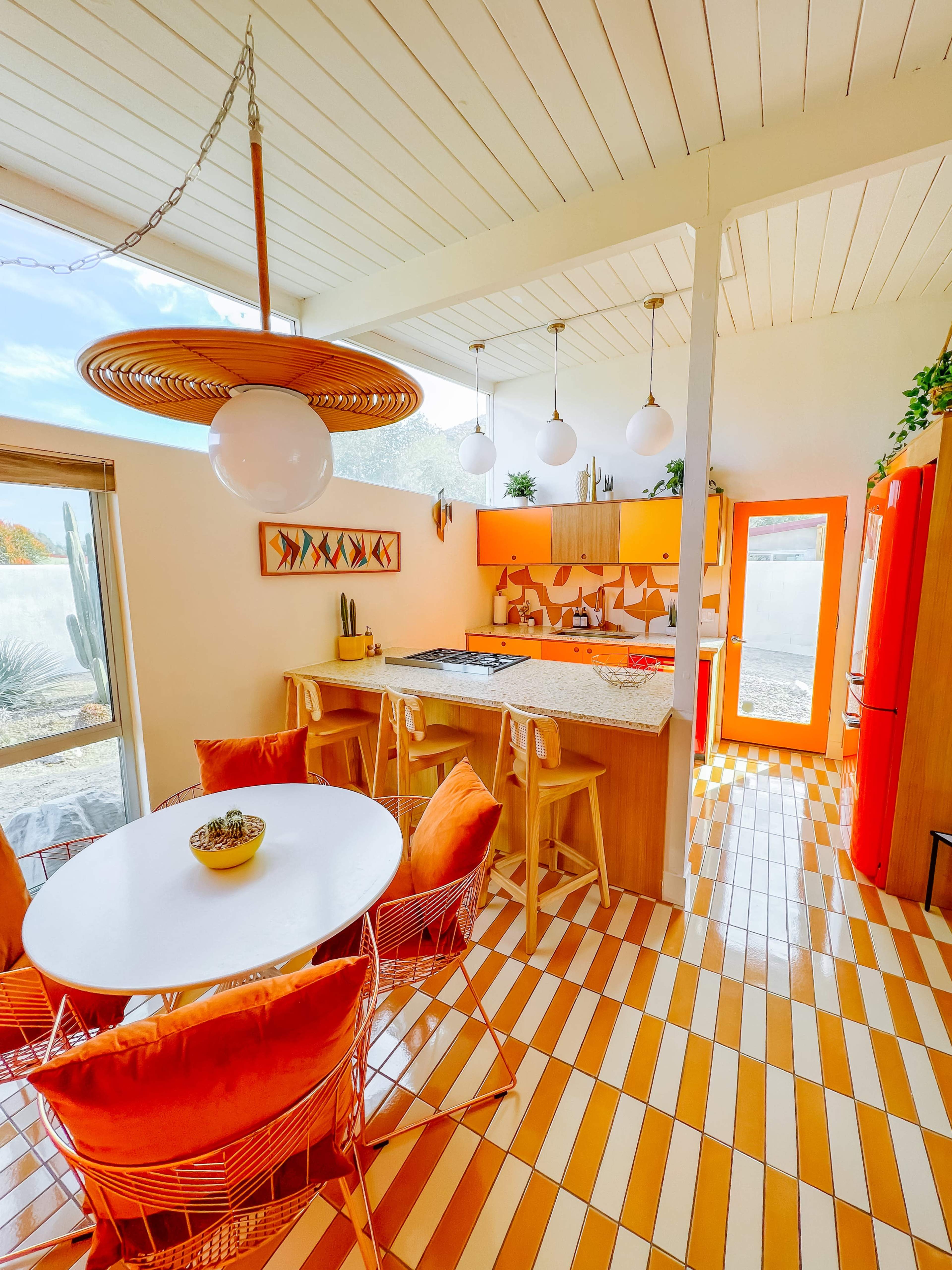 The image shows a brightly colored kitchen with orange cabinetry, a round white dining table surrounded by orange chairs, and a tiled floor in alternating stripes of orange and white.