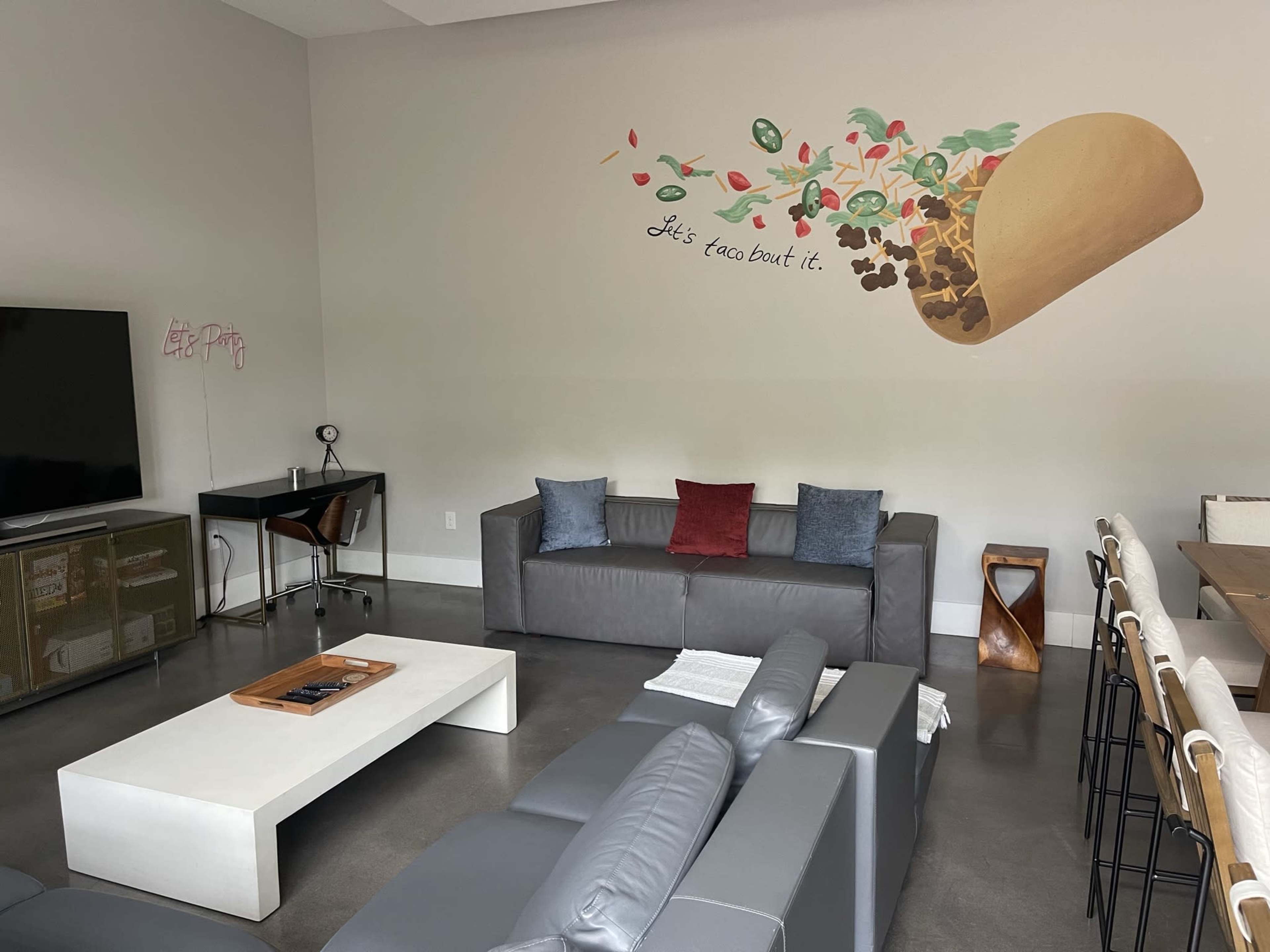 A modern living space with a large mural of a taco and the phrase "Let's taco 'bout it" on the wall, along with gray sofas, a coffee table, and a dining area.