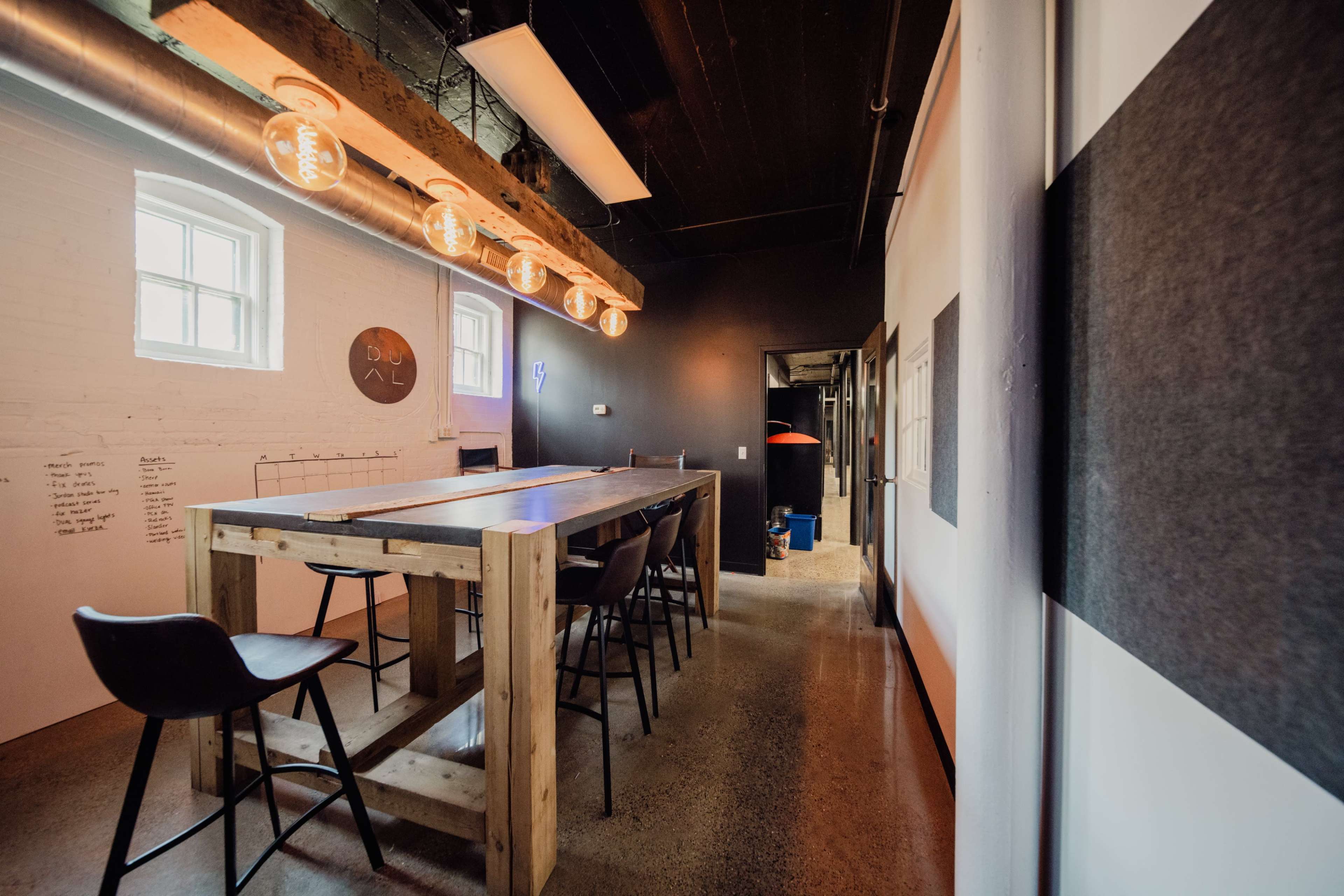 The image shows a modern, minimalist workspace featuring a large wooden table surrounded by high chairs, with industrial light fixtures hanging above.