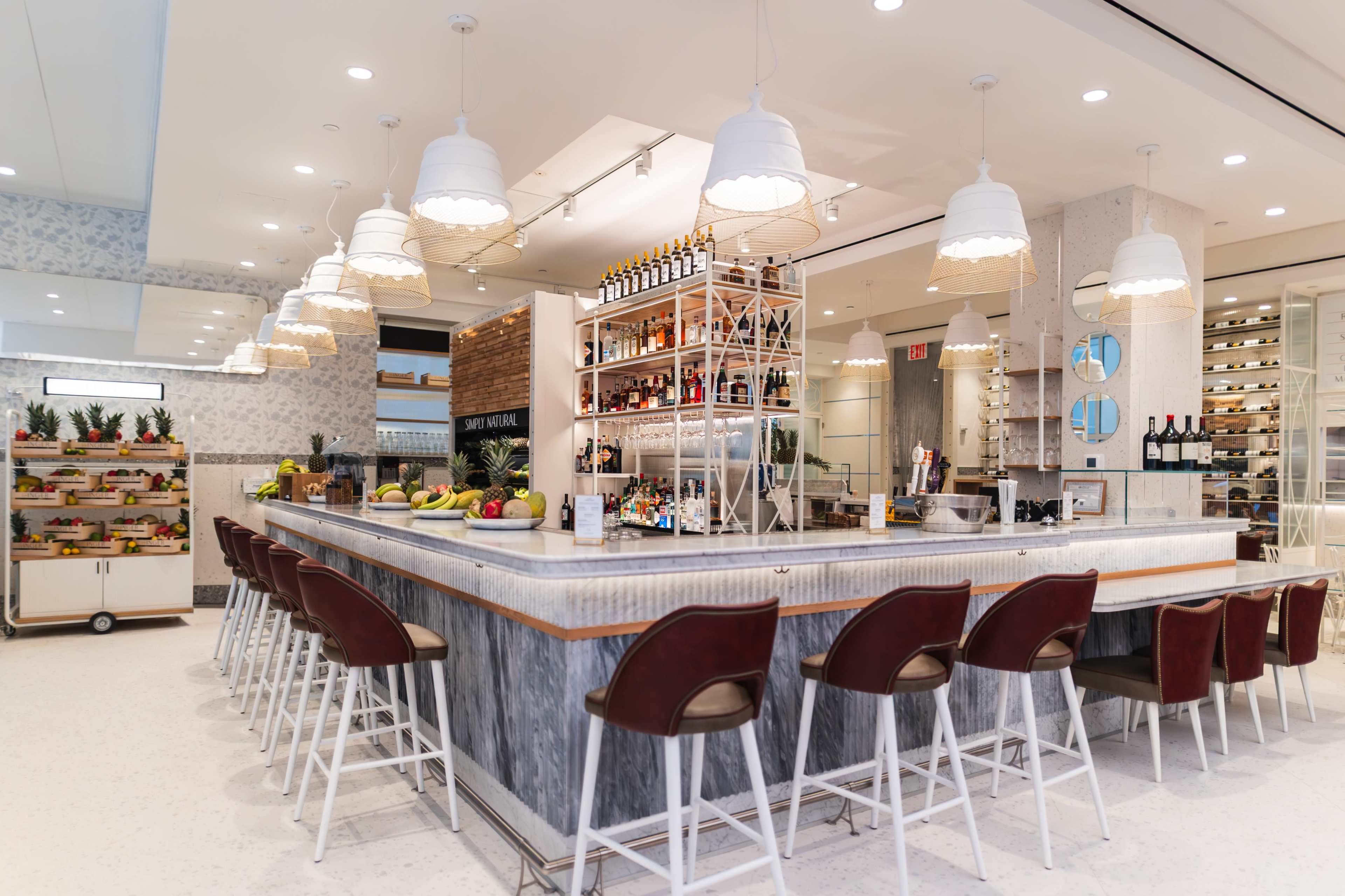 The image shows a modern bar area with a spacious counter, high stools, and shelves displaying various bottles of alcohol, all set in a brightly lit environment with white and grey decor.