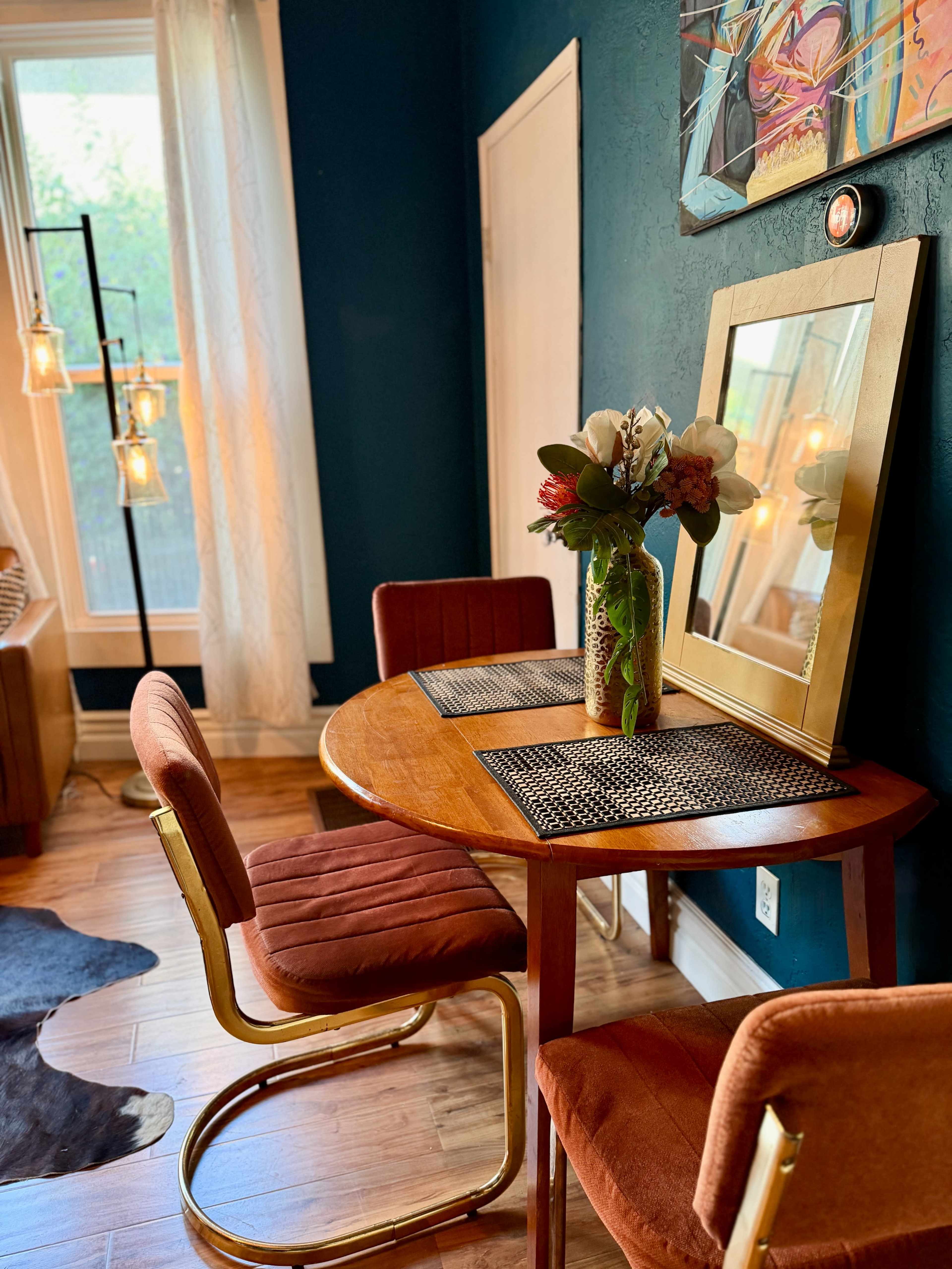 A small dining area features a round wooden table with two chairs, a vase of flowers, and a mirror, all set against a dark blue wall.