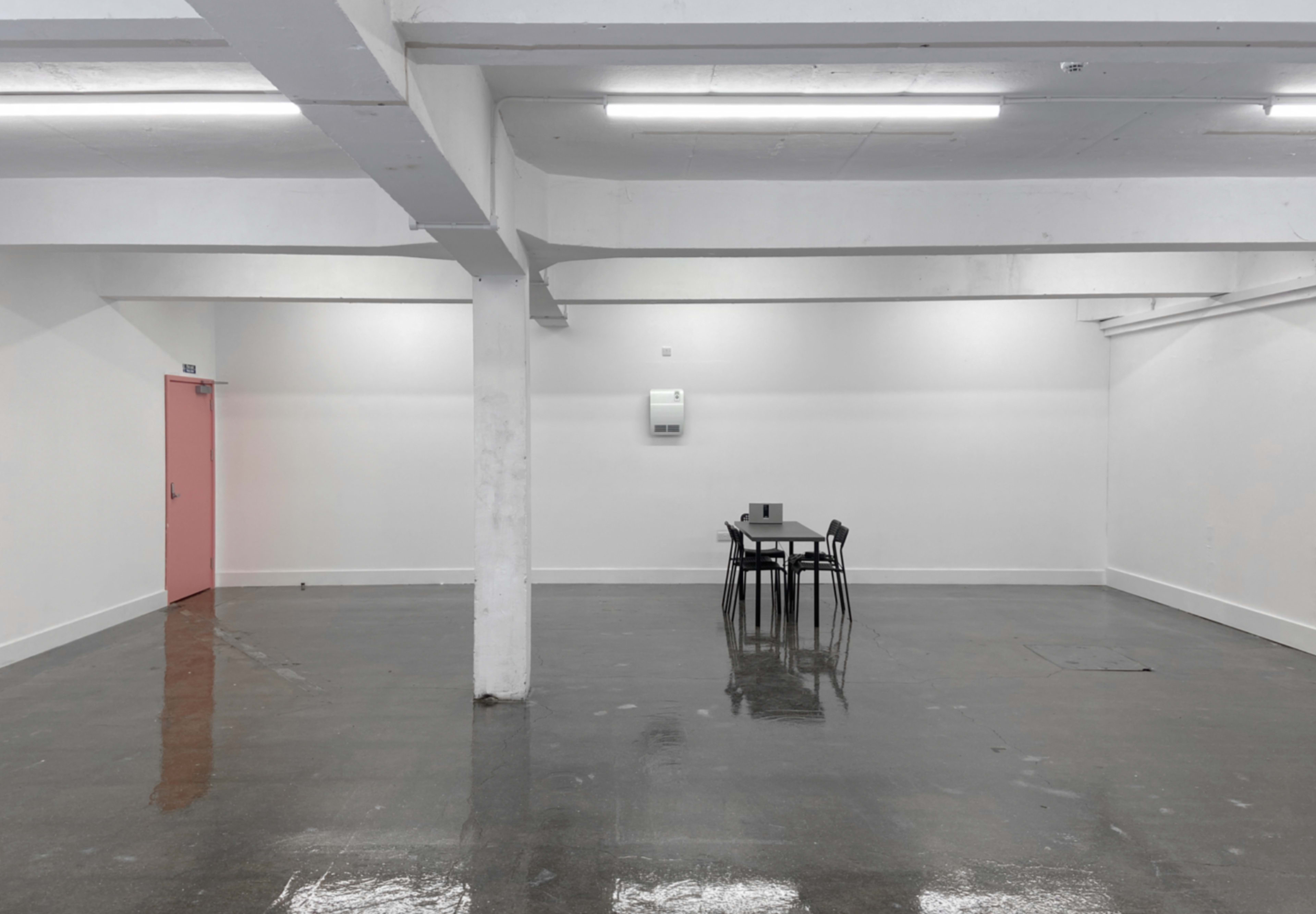 A minimalist interior space features a small table with four black chairs in the center, surrounded by white walls and a glossy floor.