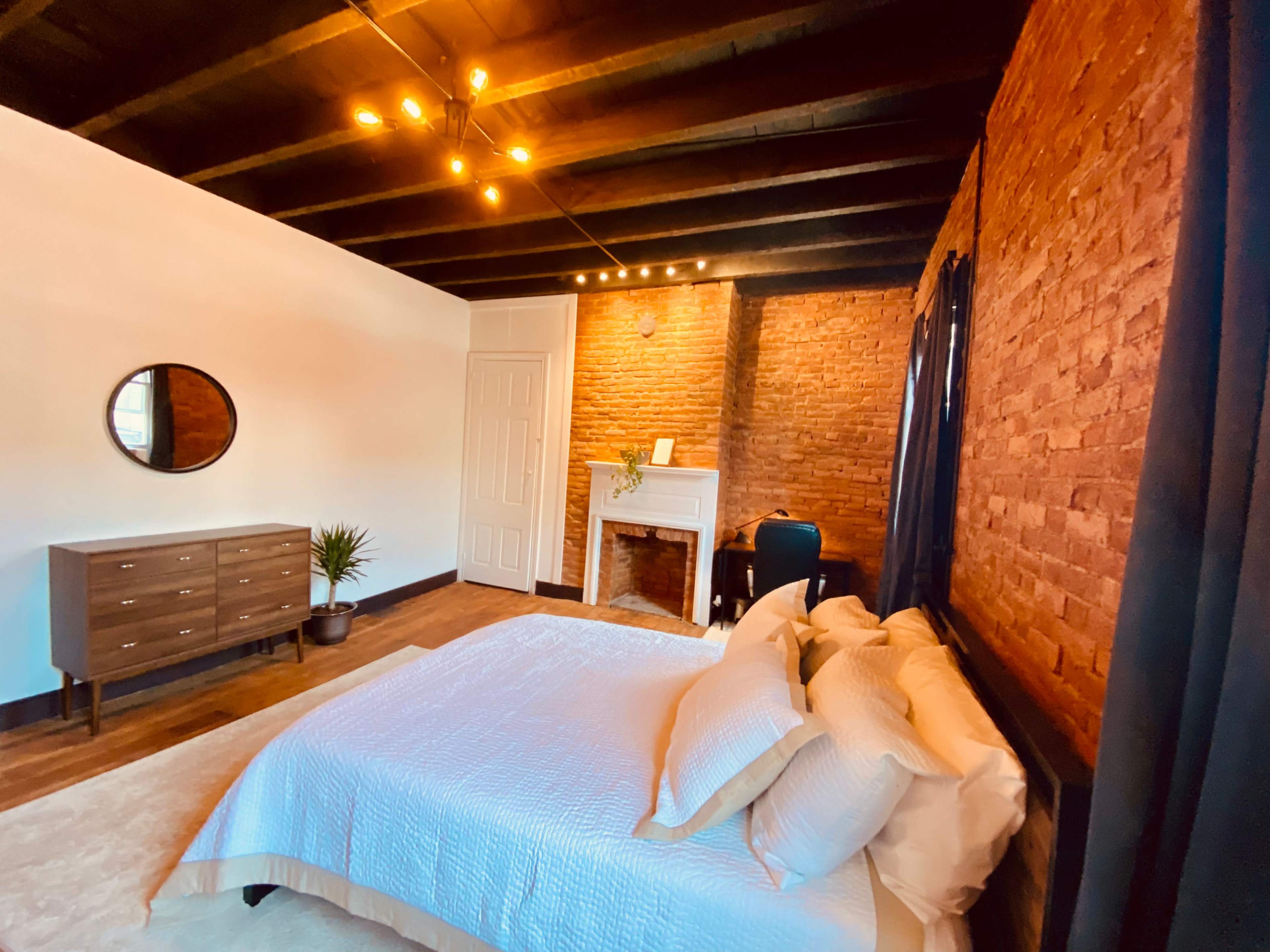 A bedroom featuring a bed with light-colored bedding, a wooden dresser, a round mirror, and exposed brick walls.