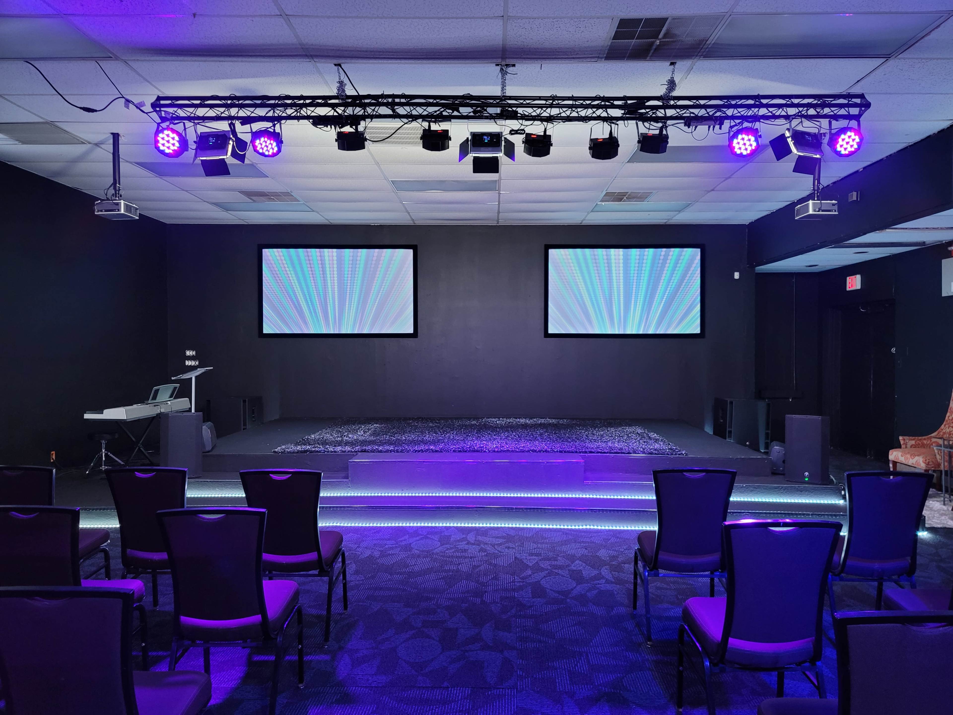A stage setup with two screens, a sound system, and rows of chairs arranged in front of it, all illuminated by purple lighting.