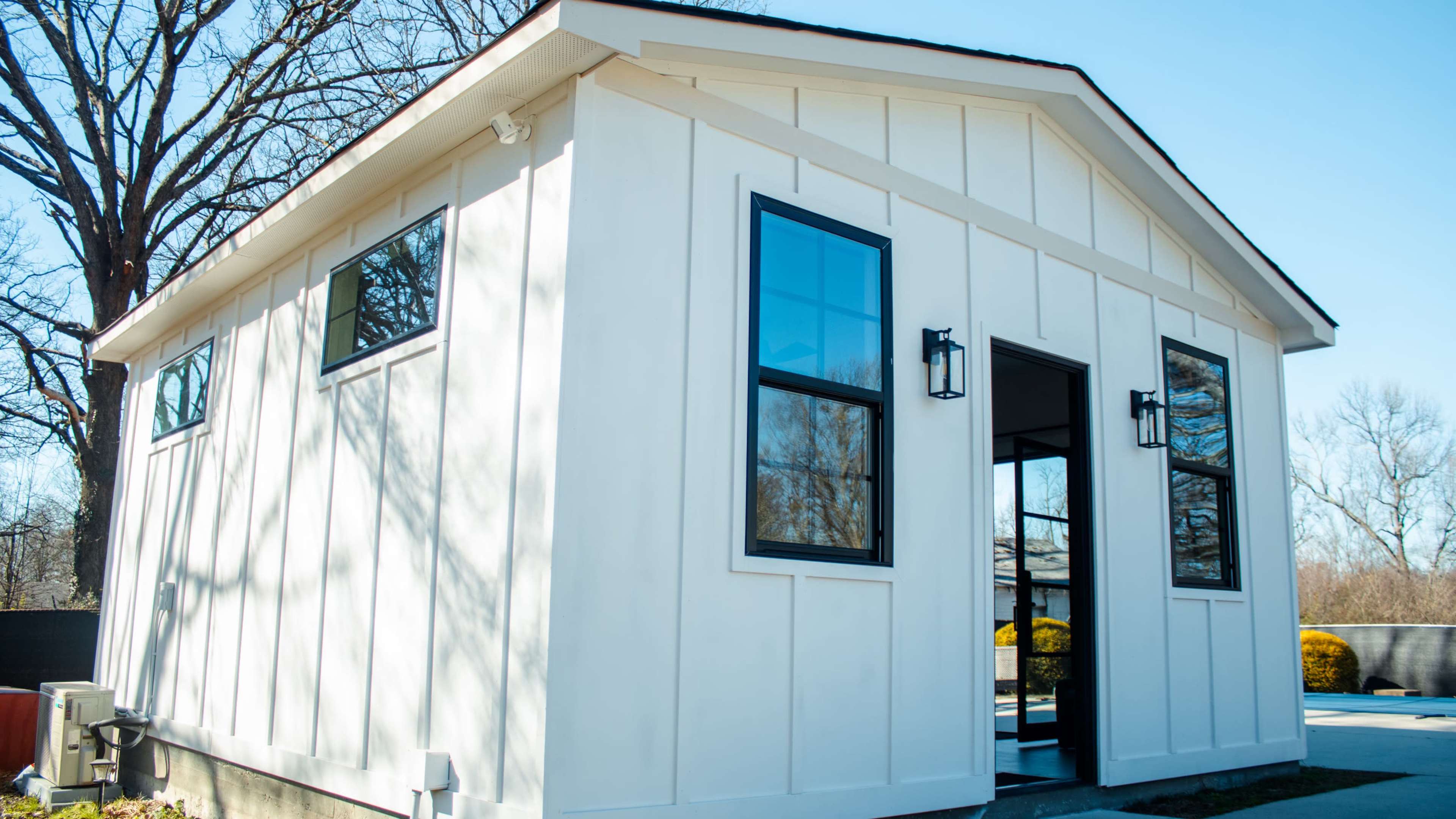 The image shows a small, modern house with a gabled roof, white exterior walls, and large windows, situated in a landscaped area.