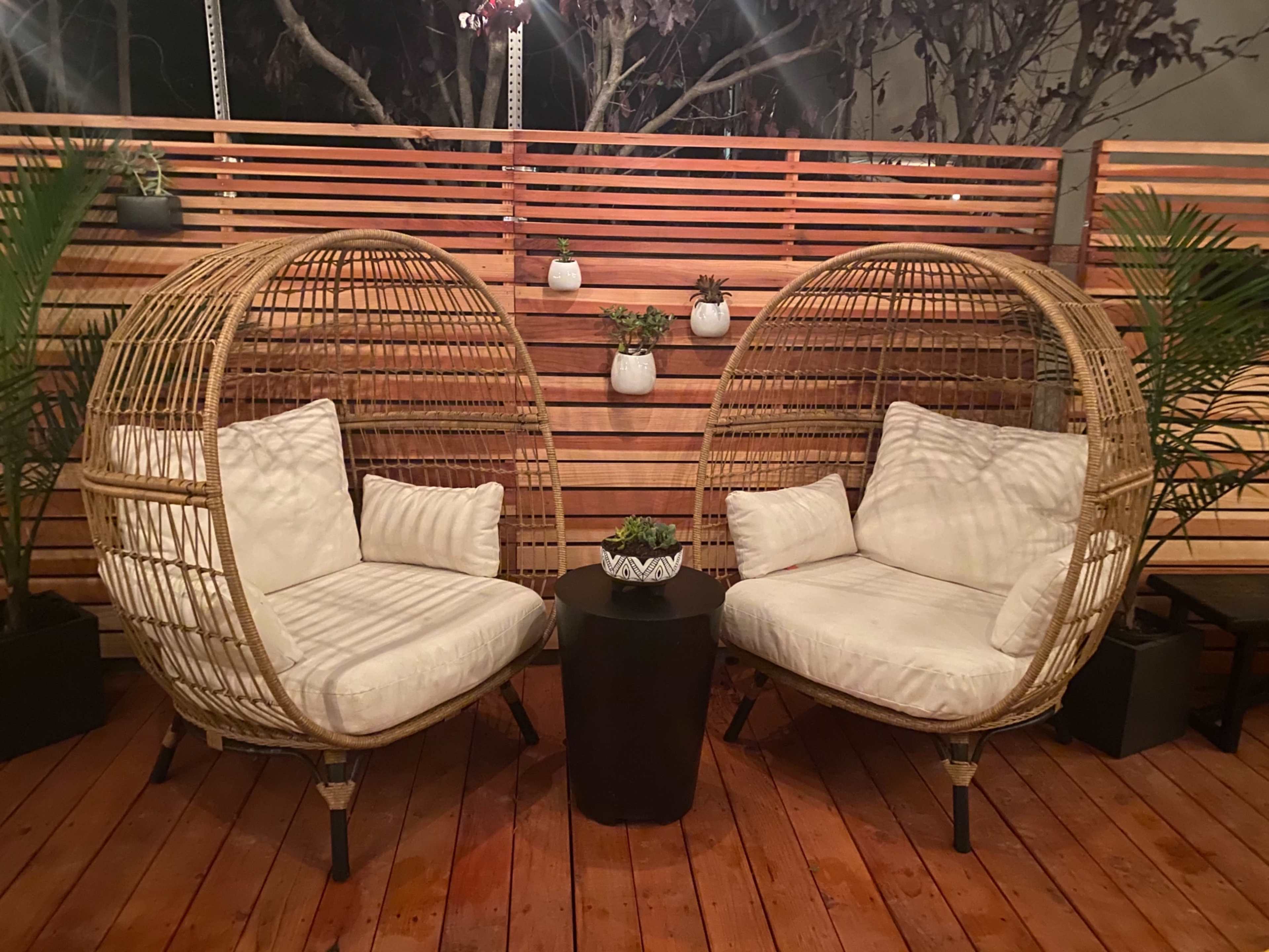 Two round wicker chairs with cushions are situated facing each other on a wooden deck, accompanied by a small black table and potted plants in the background.