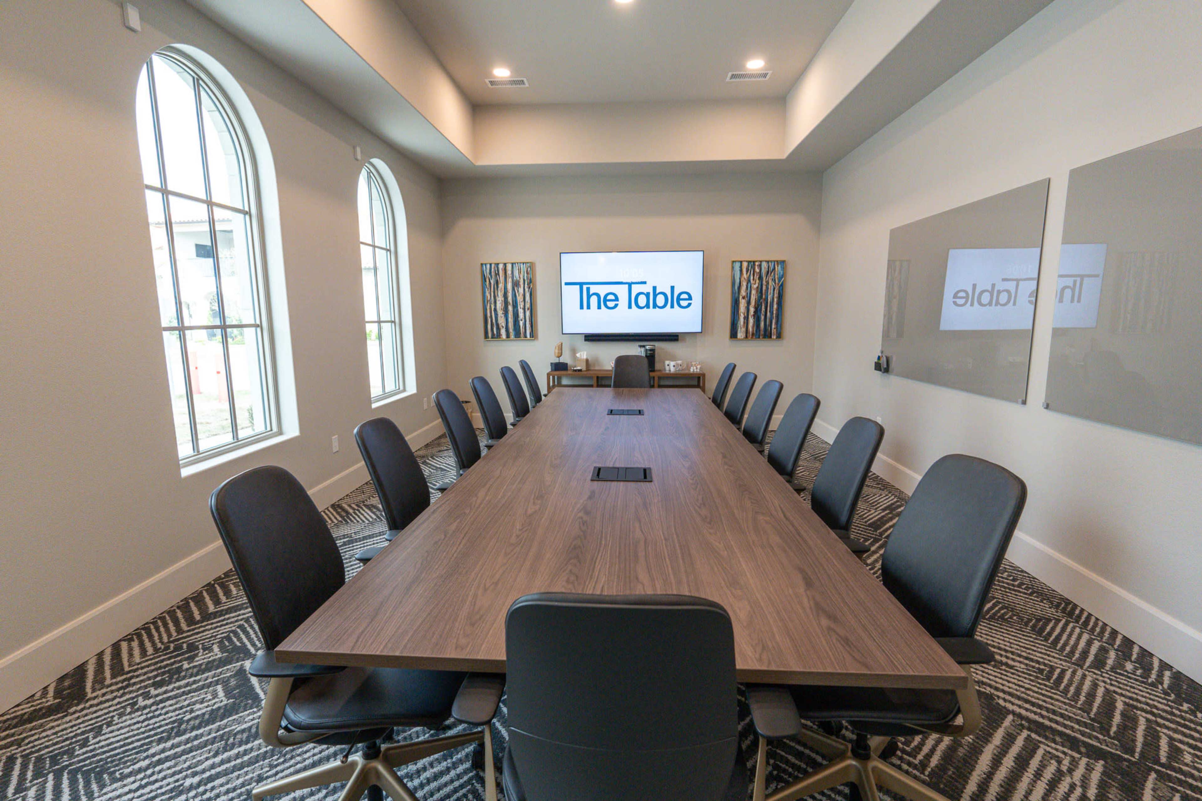 A long conference table with black chairs is set up in a well-lit room featuring large windows and two displays on the walls.