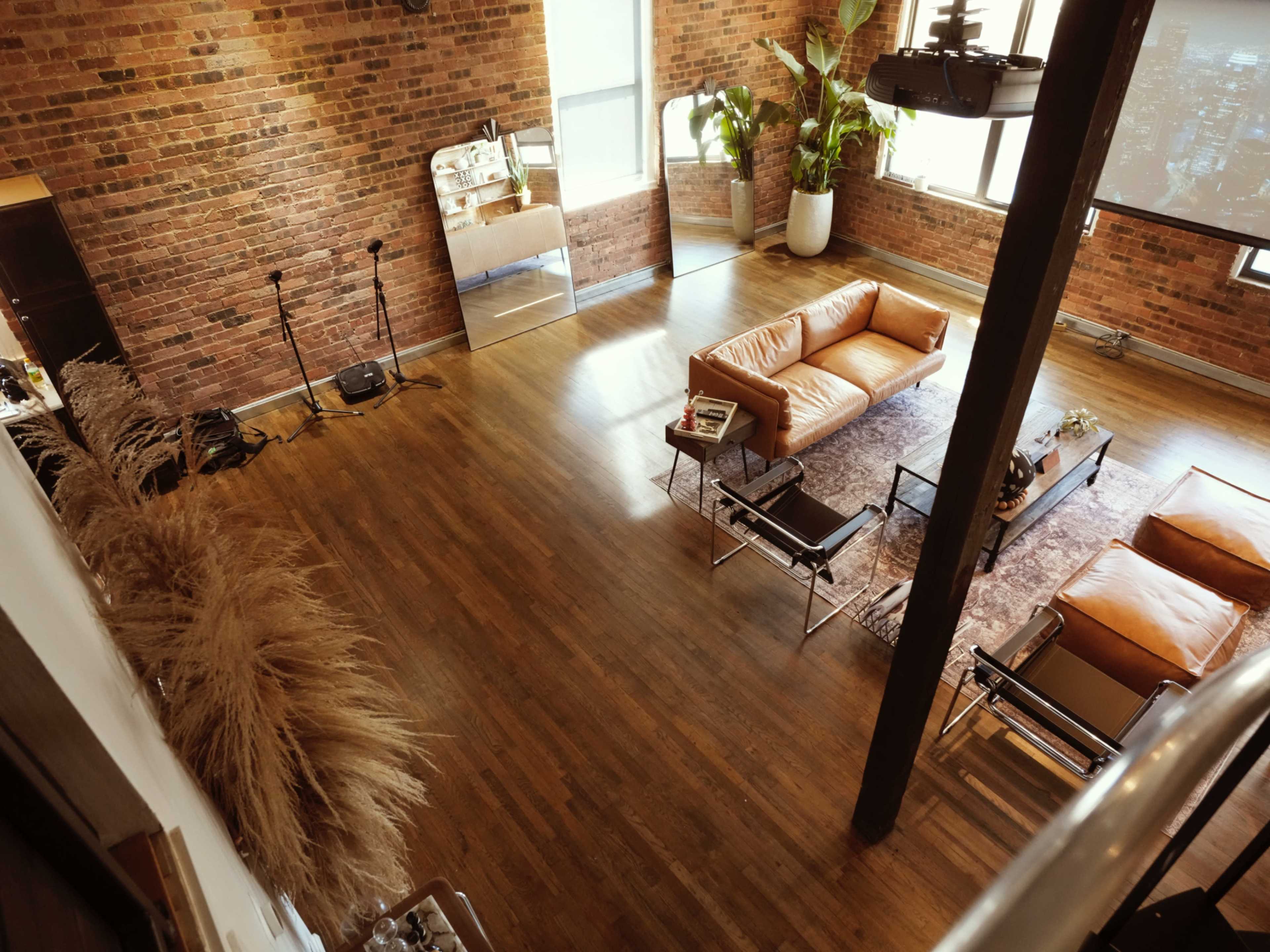 The image depicts a spacious room with exposed brick walls, hardwood flooring, and various seating arrangements, including a large orange sofa and plants.