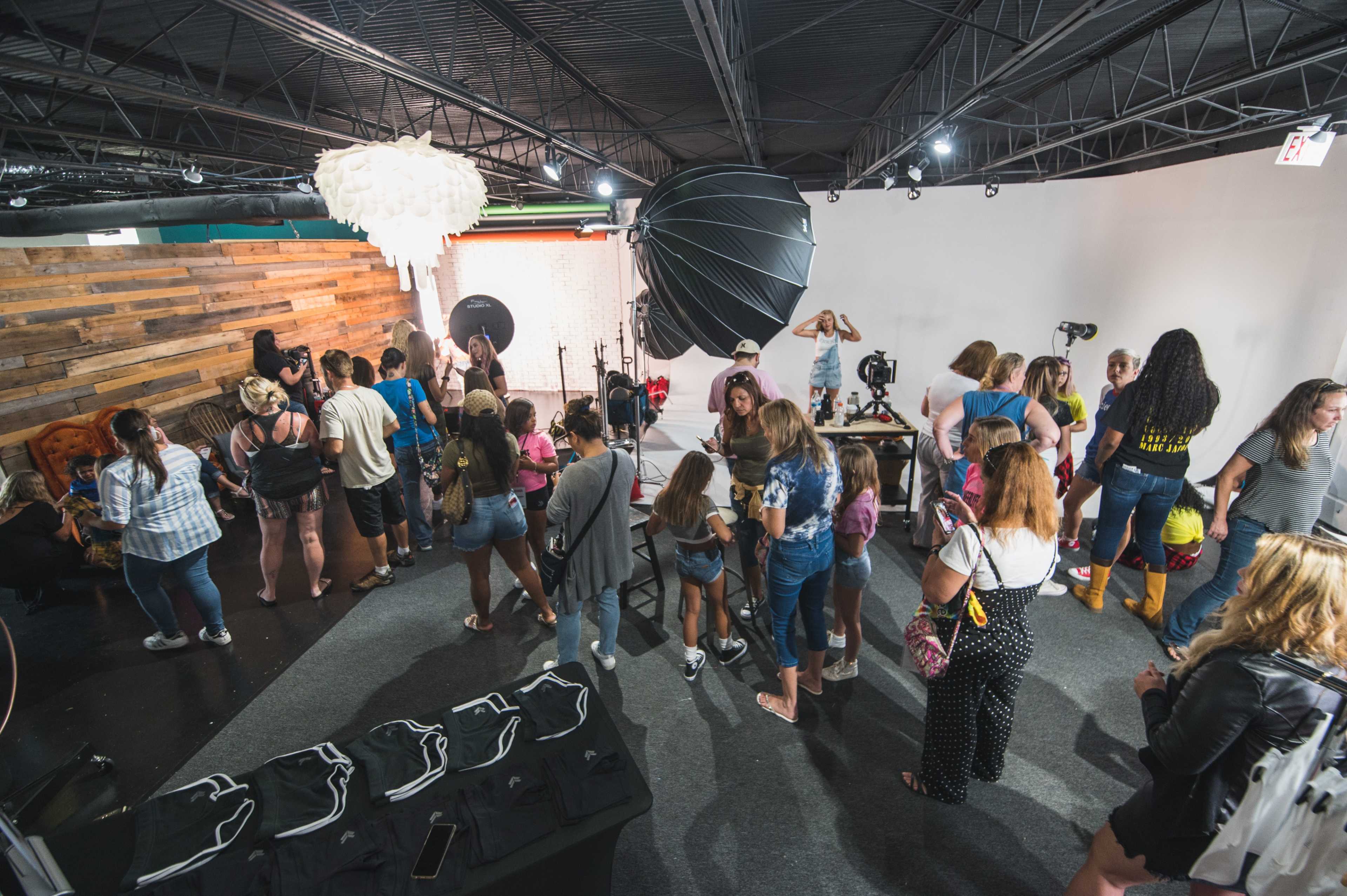 A crowded photography studio features numerous people engaged in various activities, with large backdrop setups and lighting equipment visible.