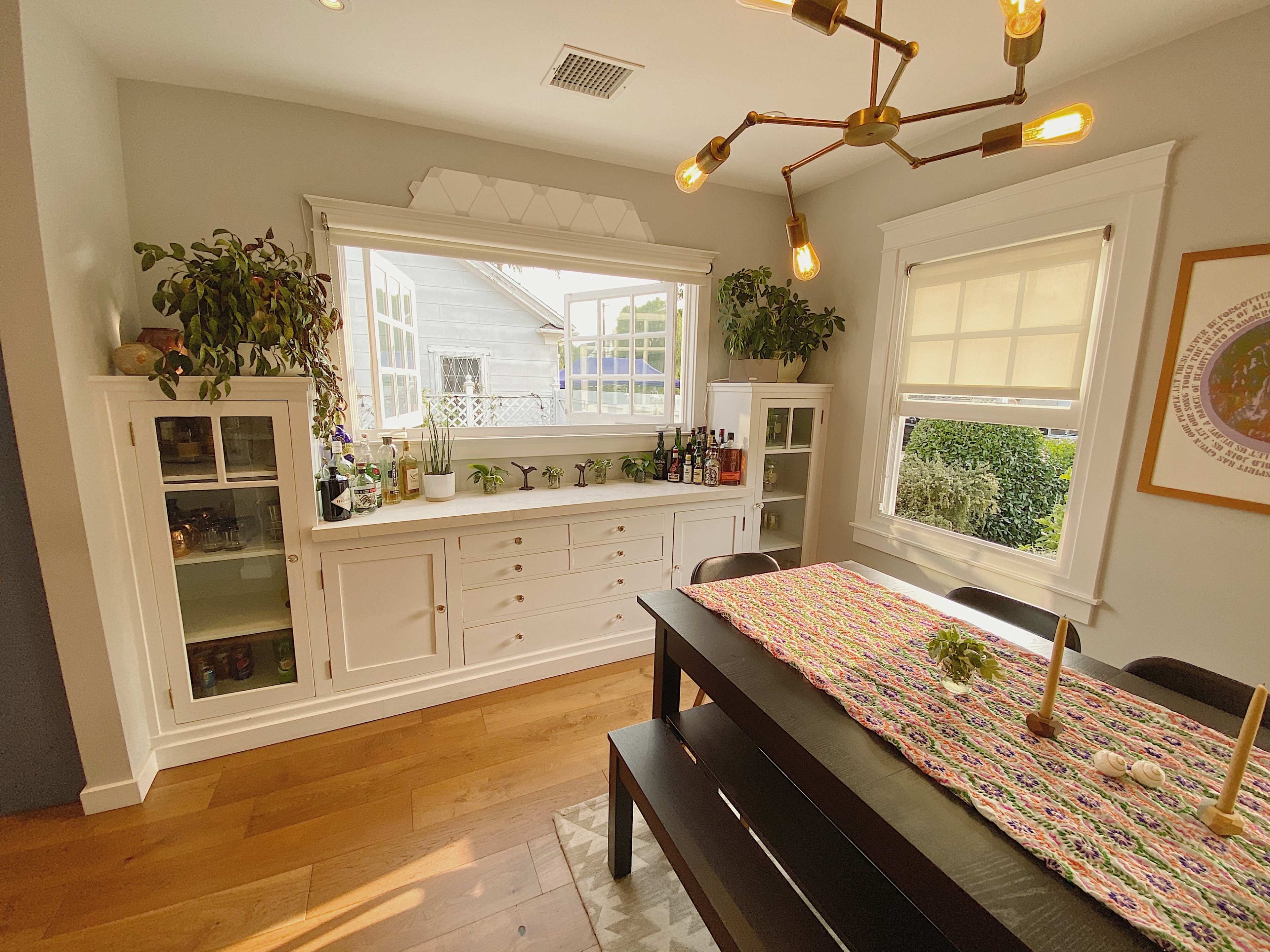A bright dining area with a black table, a colorful table runner, and a white sideboard featuring plants and bottles underneath a window.