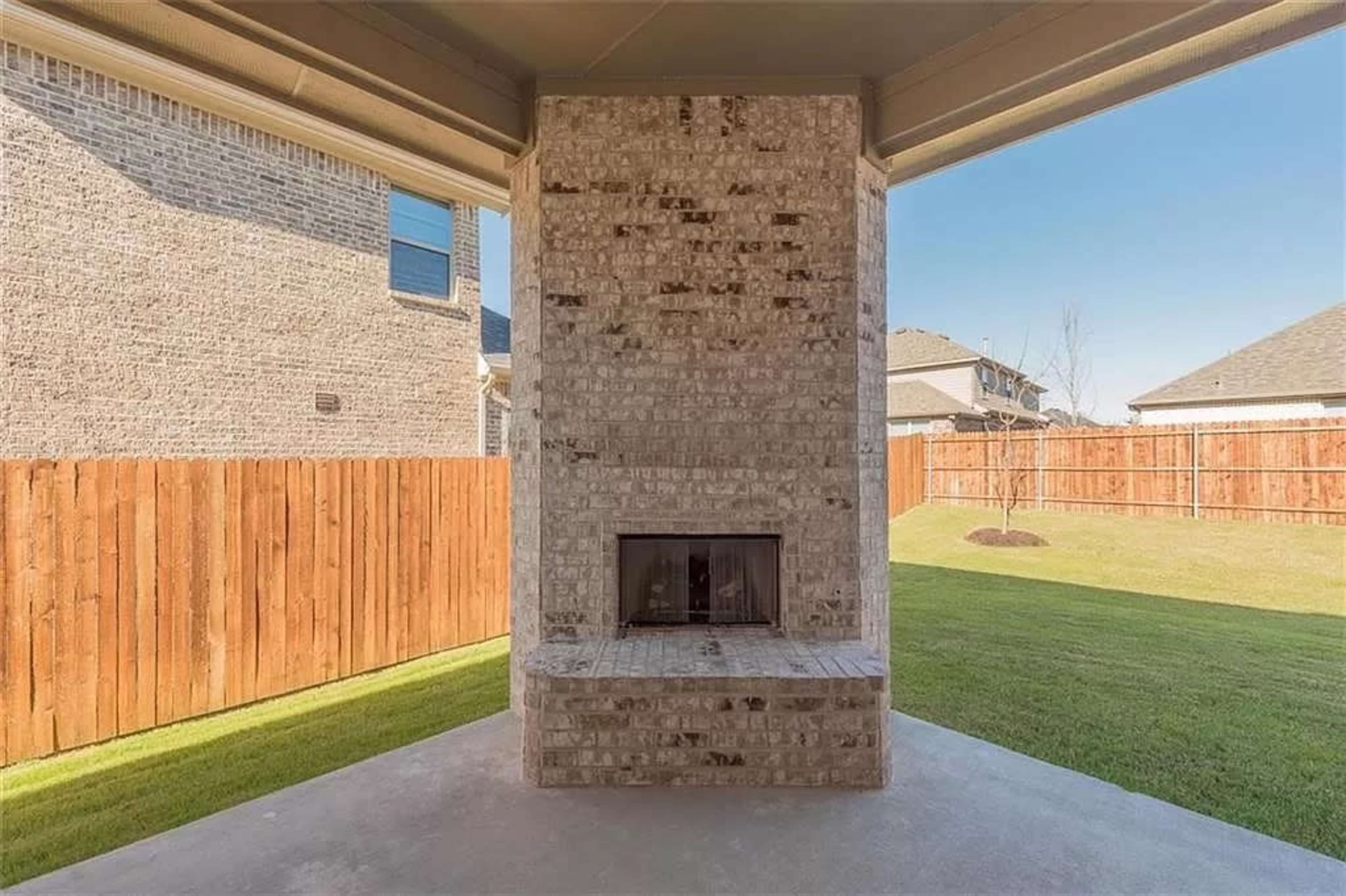 A brick outdoor fireplace situated on a concrete patio, surrounded by a grassy yard and a wooden fence.