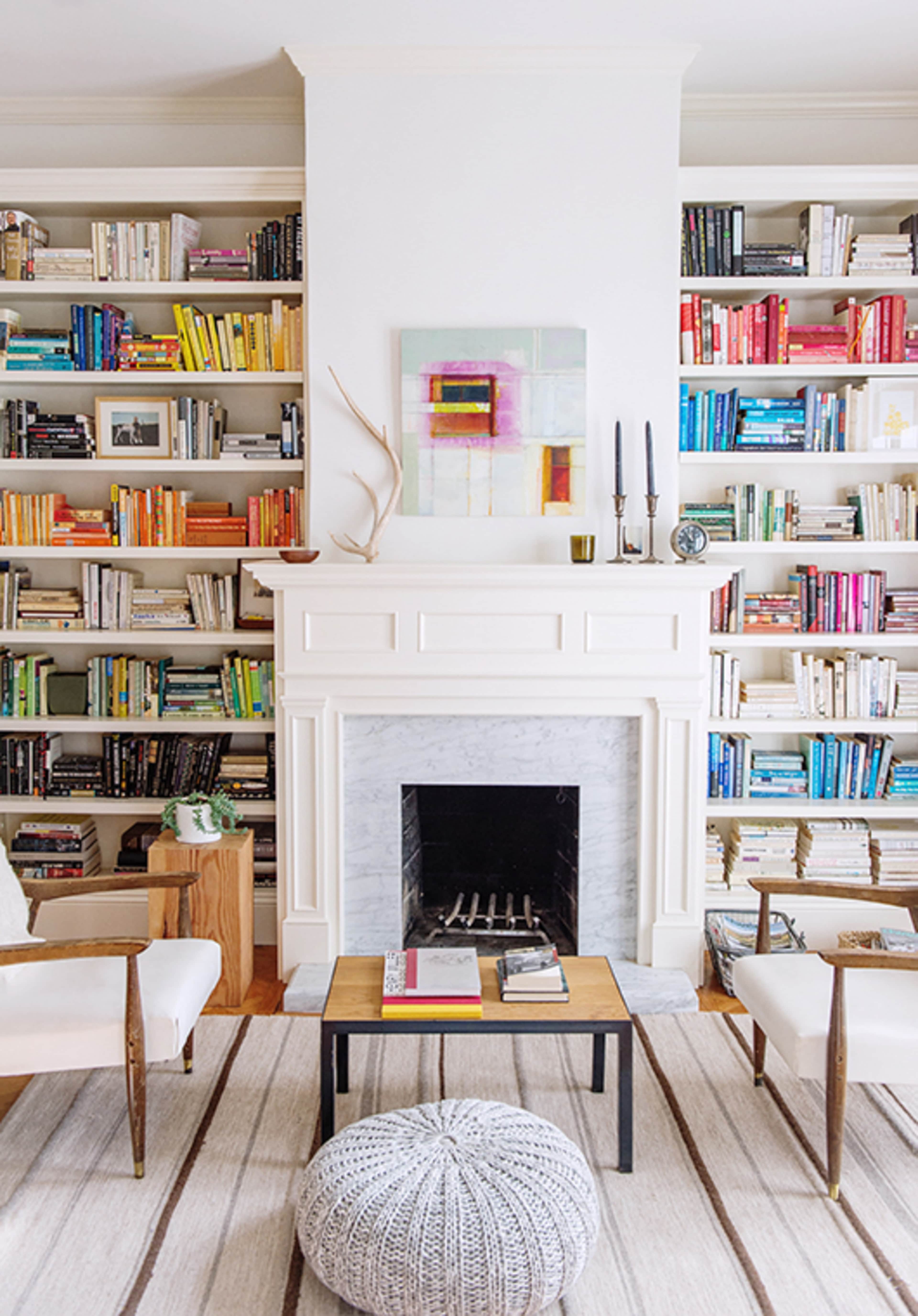 A living room with a white fireplace flanked by bookshelves filled with color-coordinated books, a coffee table in front, and two armchairs on either side.