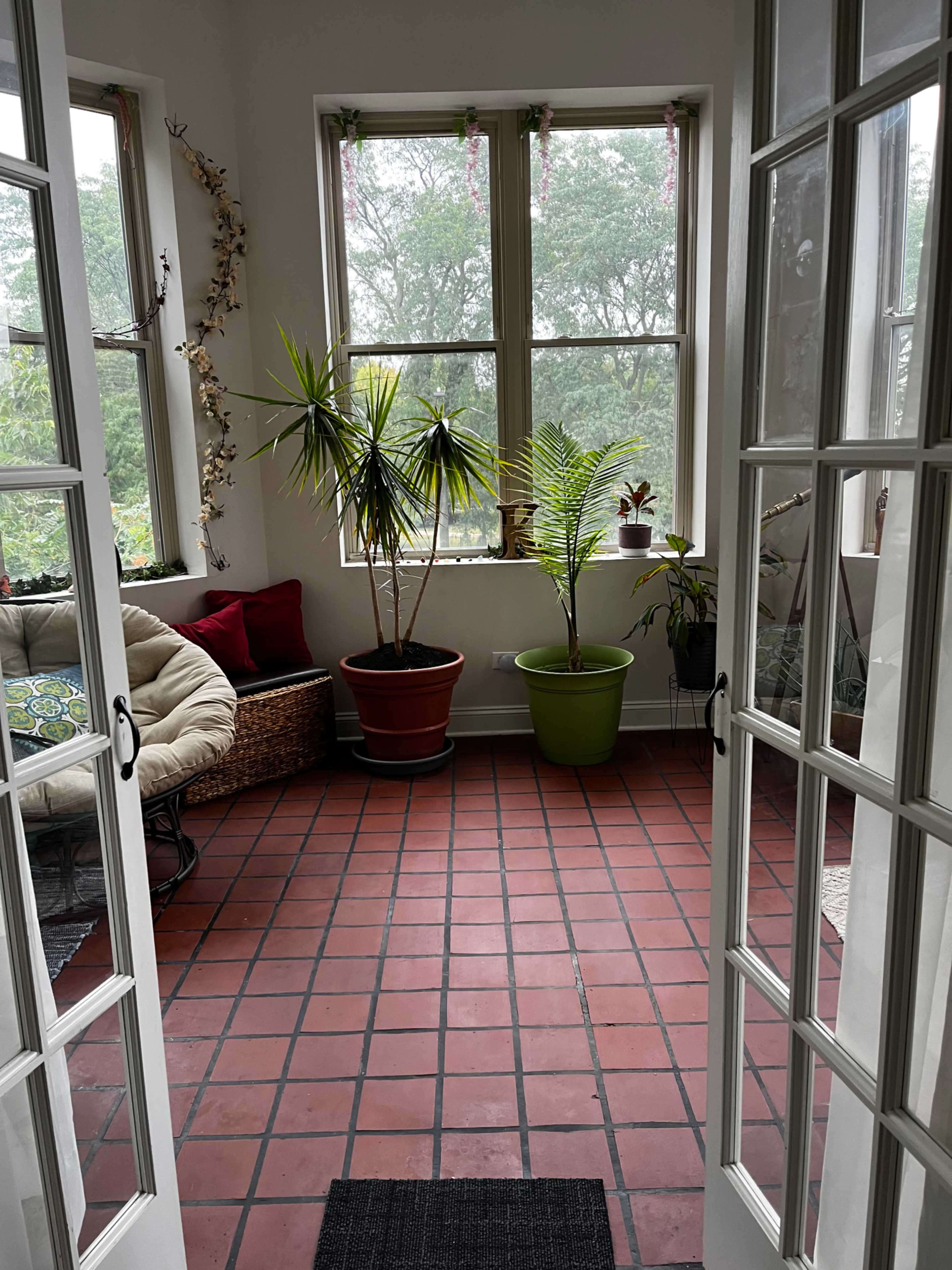 The image shows a sunroom with red tile flooring, two potted plants, a hanging chair, and large windows allowing light to enter.