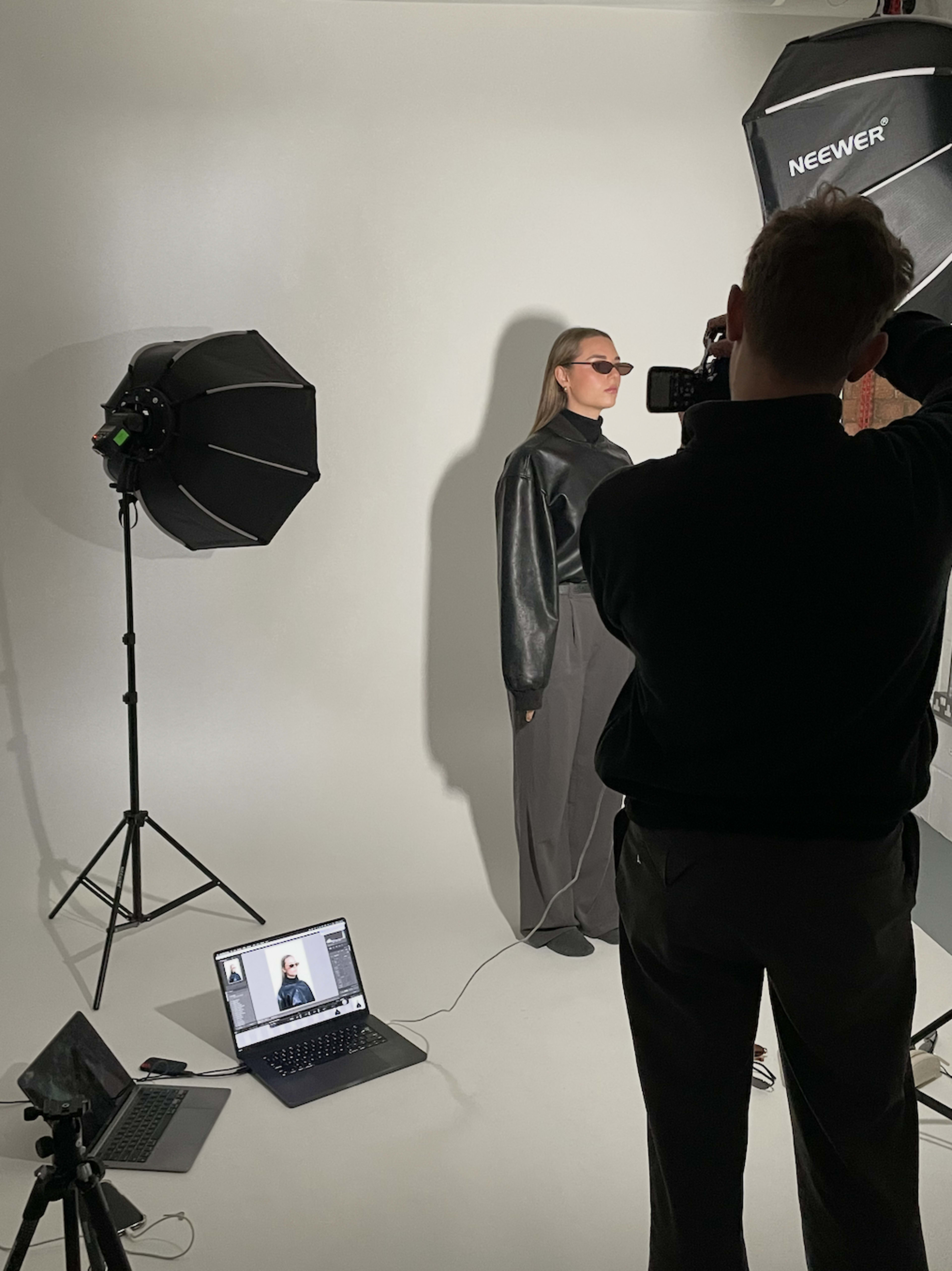 A photographer is capturing images of a model dressed in a black outfit against a neutral backdrop, with studio lighting and a laptop displaying the camera feed positioned nearby.