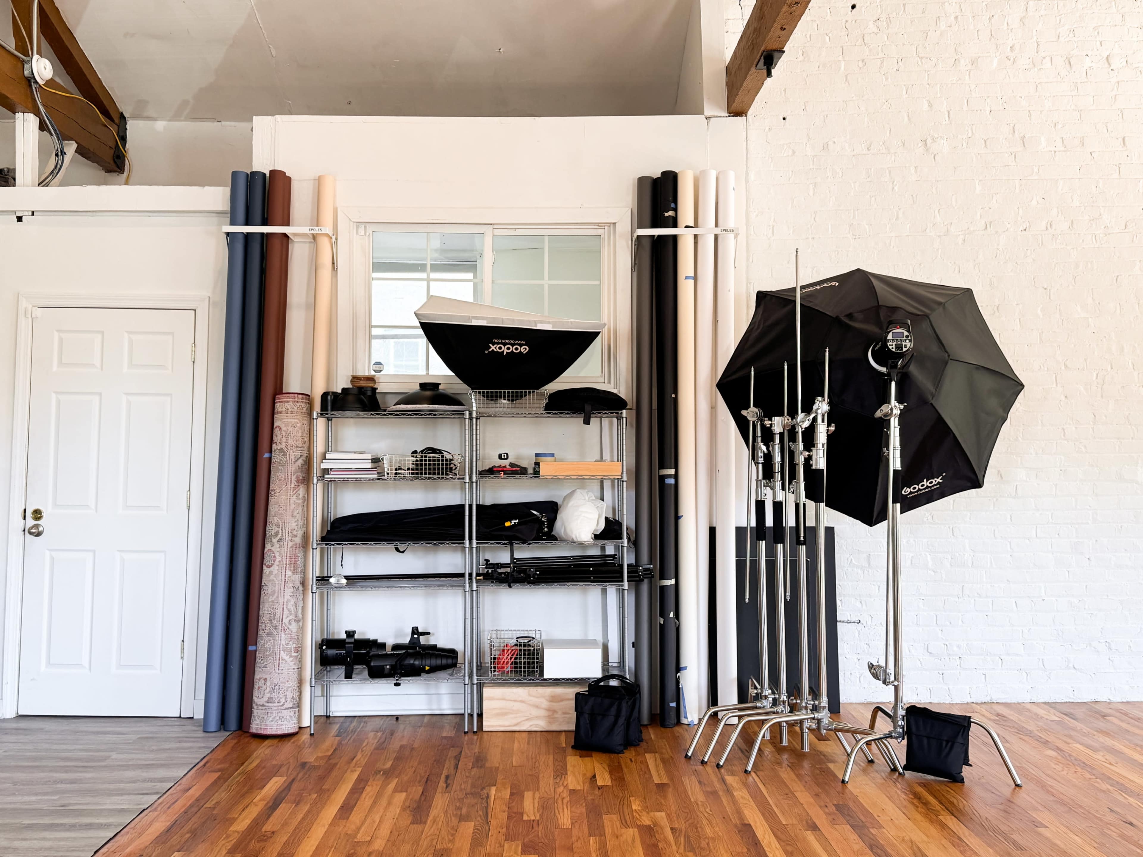 The image shows a photography studio setup with rolls of backdrop paper, lighting equipment, and various accessories organized on shelves and stands against a white wall.