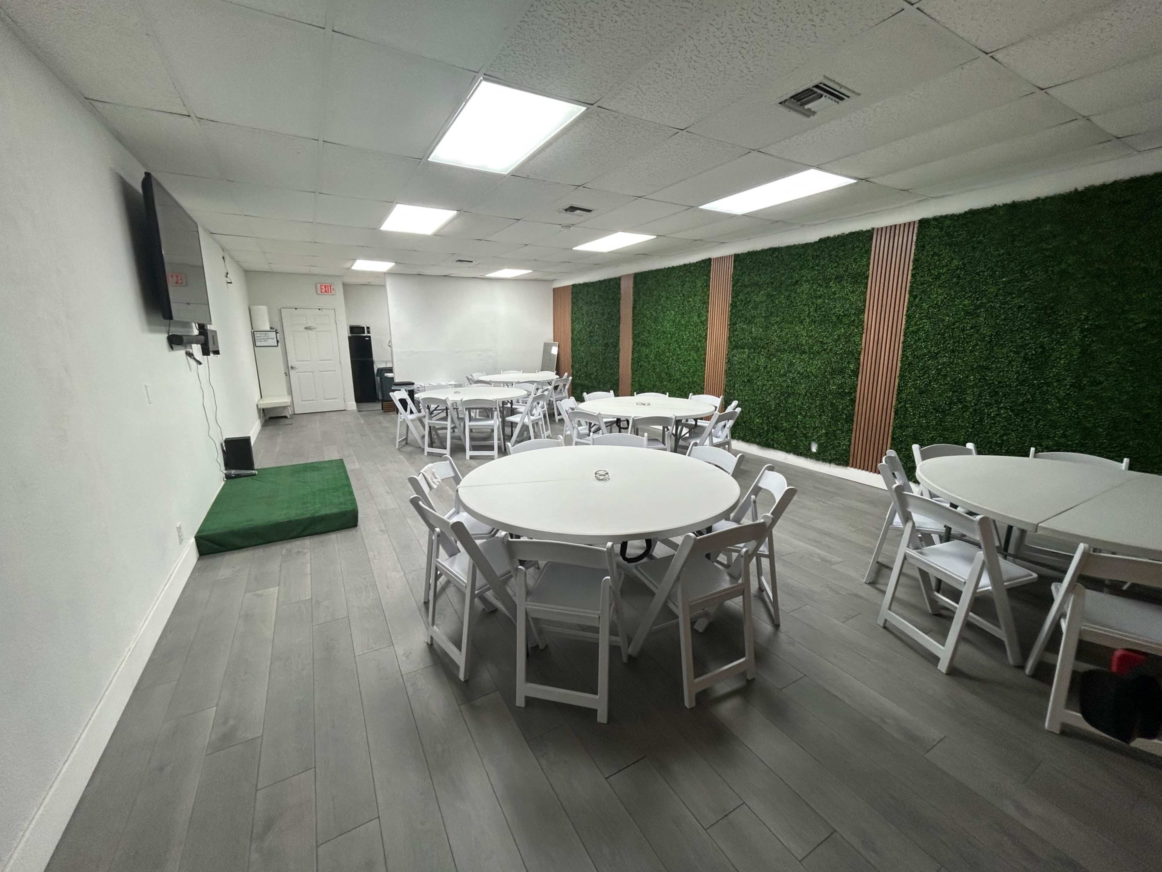 The image shows a spacious room with multiple round tables and chairs, a green wall, and a television mounted on the wall.