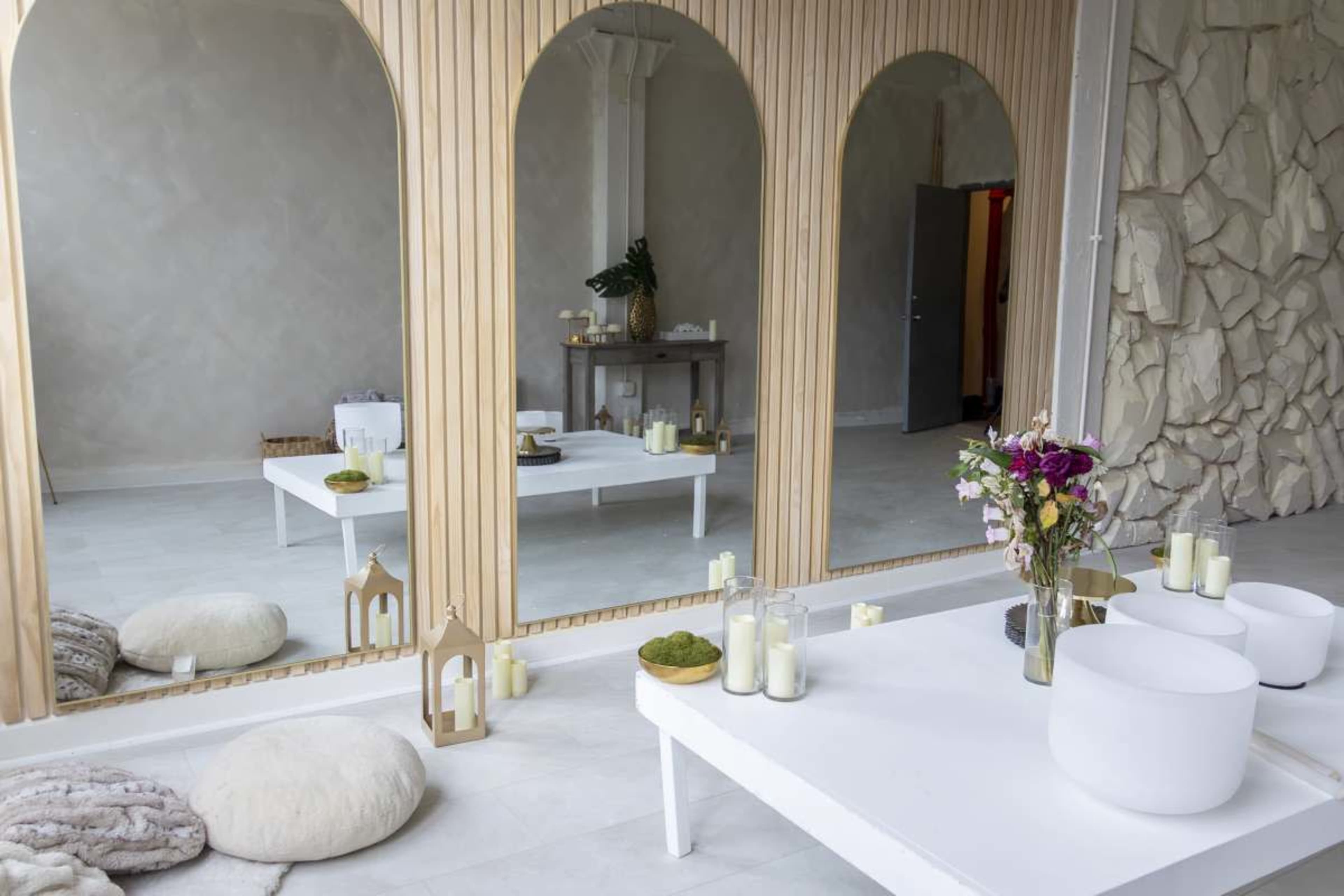 The image shows a minimalist interior space featuring three large arched mirrors, a low white coffee table adorned with decorative candles and flowers, and soft cushions on a light floor.