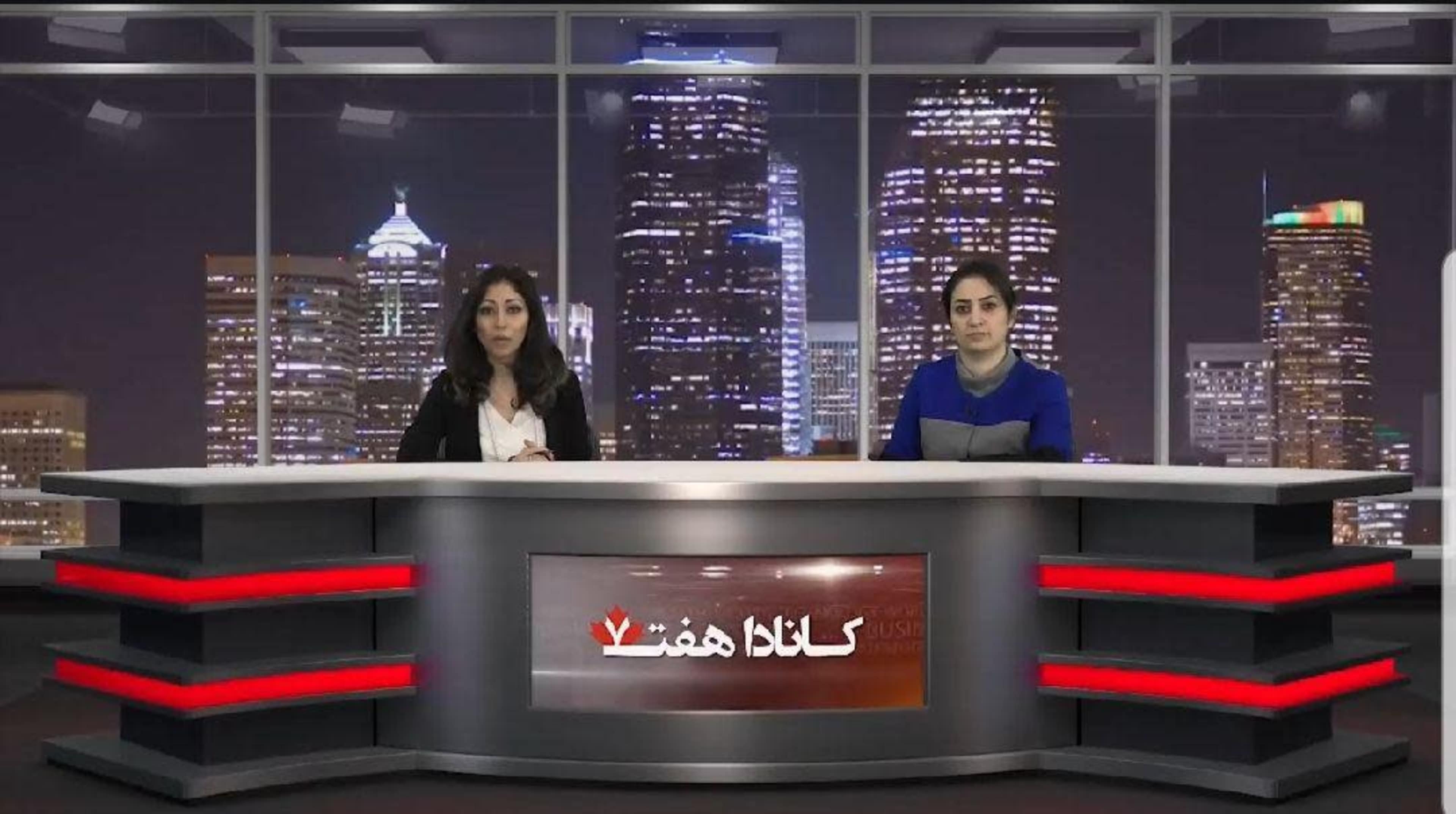 Two women are seated at a news desk in a studio, with a city skyline visible through large windows behind them.