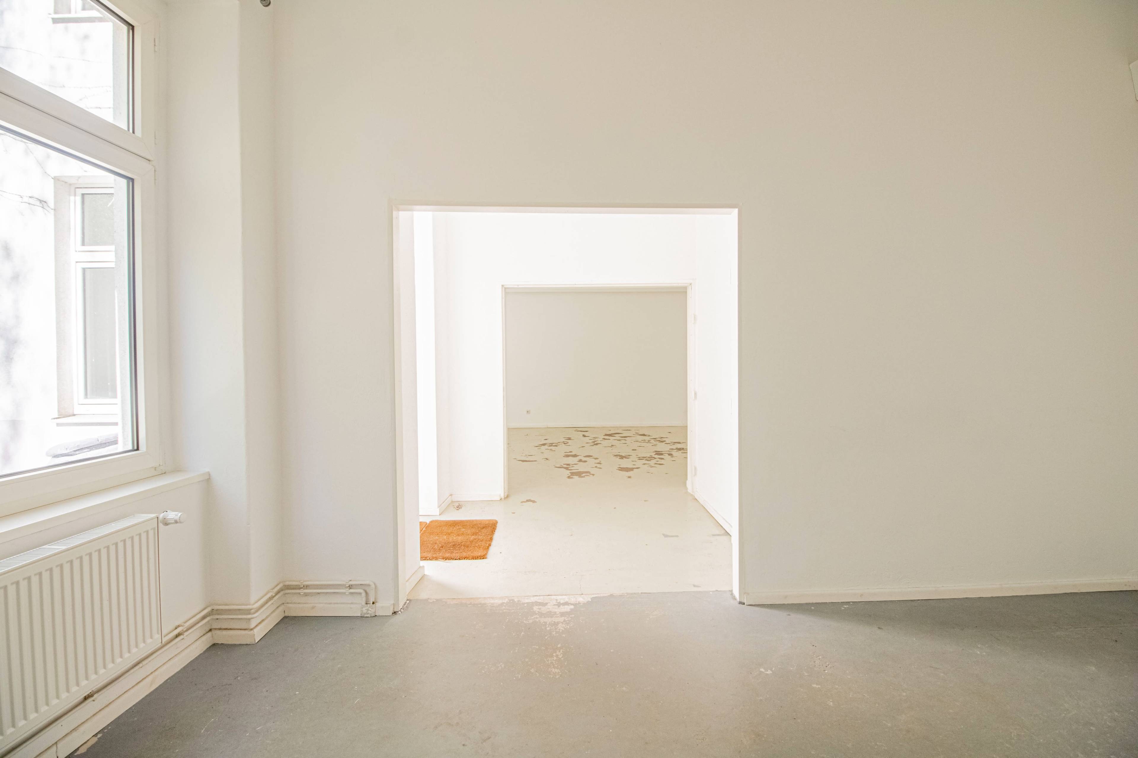 The image shows a bright, empty interior space with white walls, a bare floor, and a doorway leading to another room.