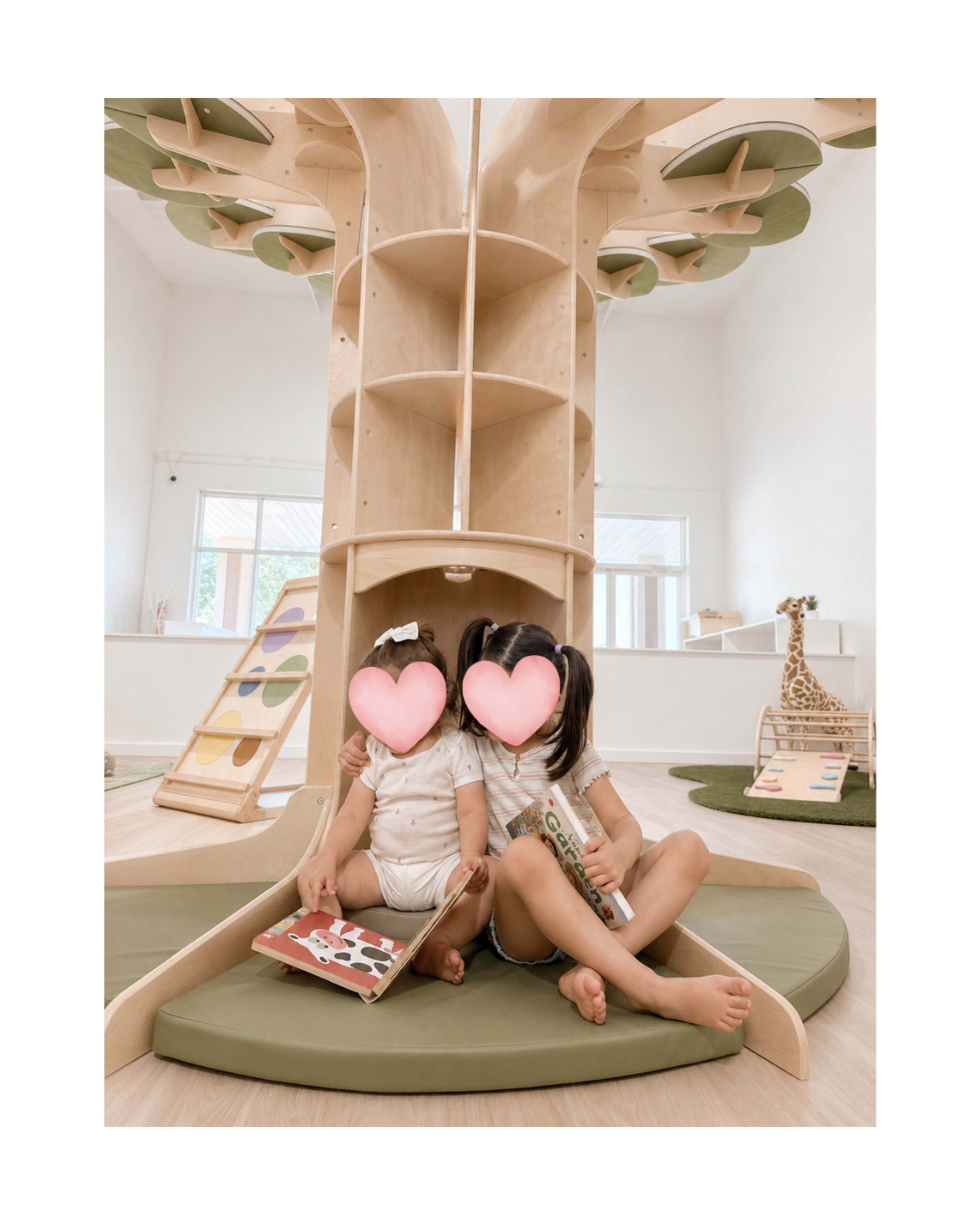 Two children sit on a green play mat inside a playroom featuring a large wooden tree structure with shelves and a slide.