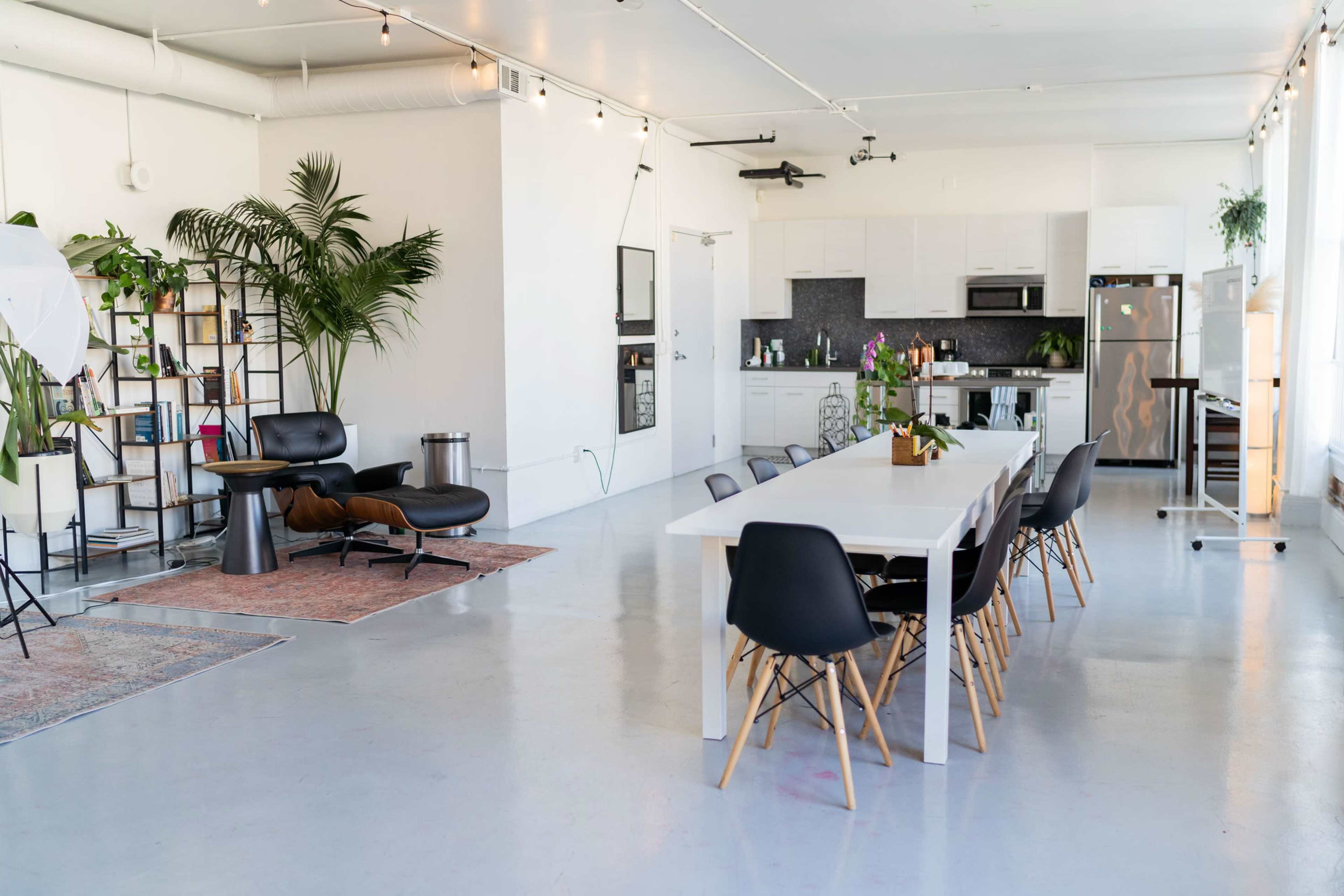 A bright, open living space featuring a dining area with a long white table, black chairs, and a lounge area with a chair and a bookshelf, all surrounded by greenery.