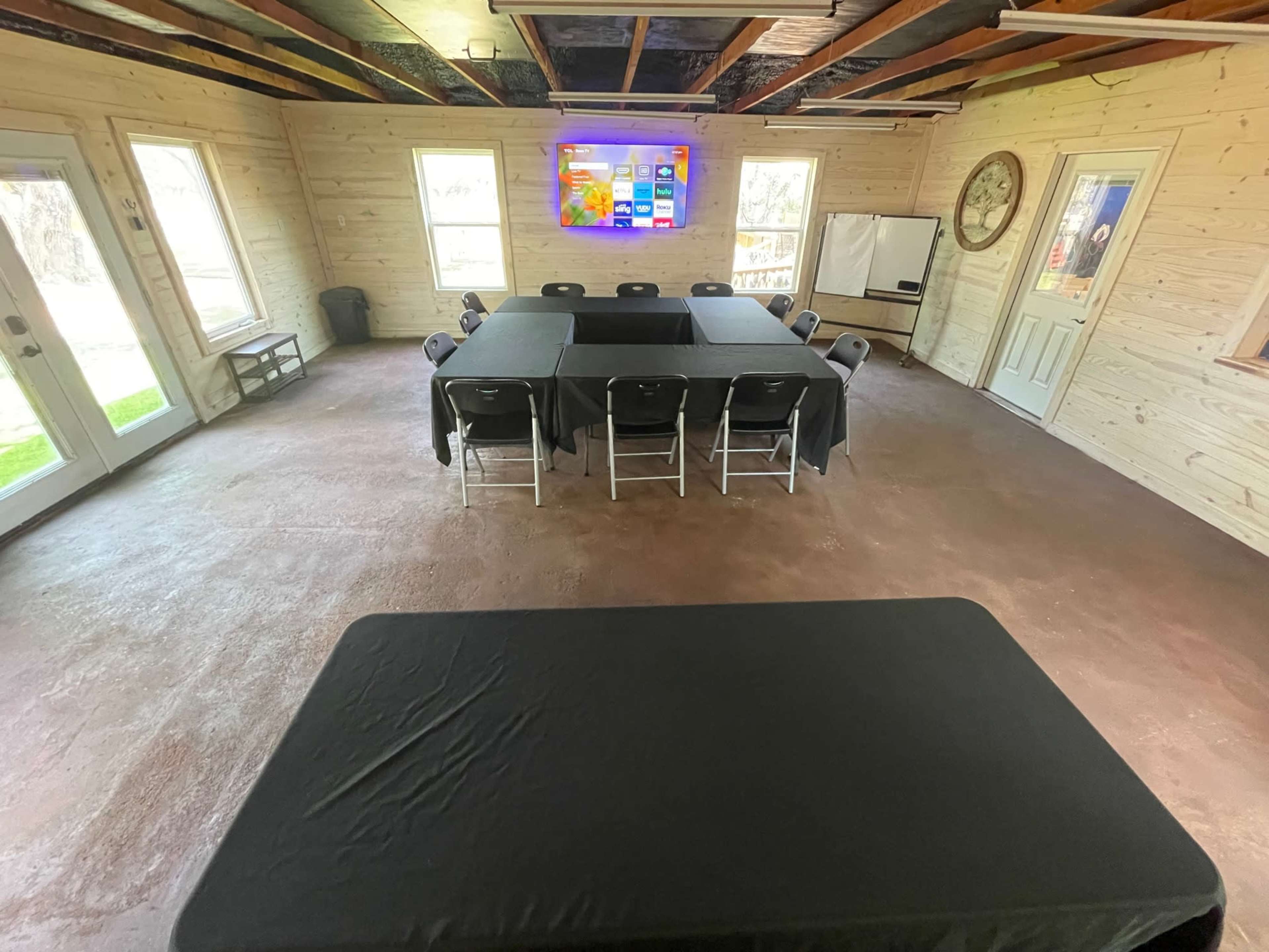 A meeting room with a central rectangular table surrounded by chairs, large windows, and a television mounted on the wall.