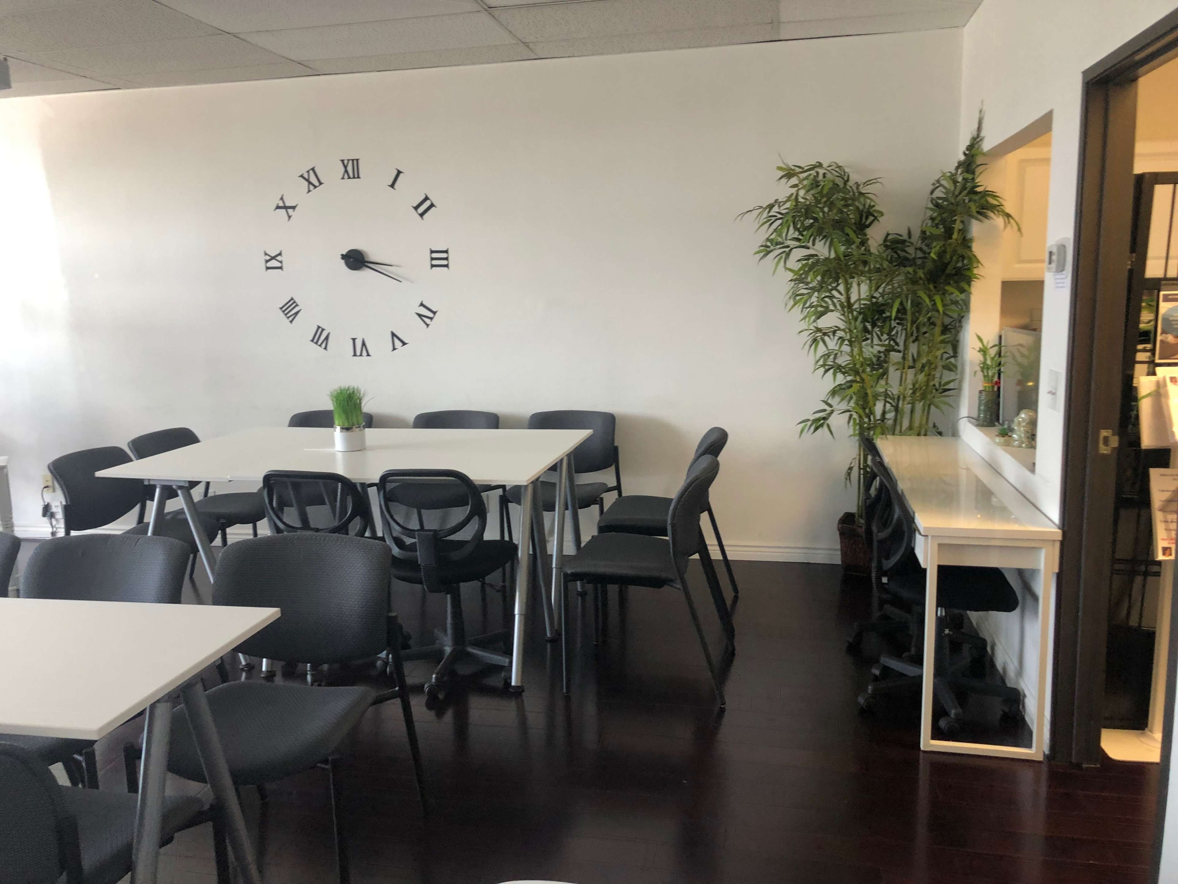 A meeting room features several white tables and black chairs, with a large wall clock and a potted plant in the corner.