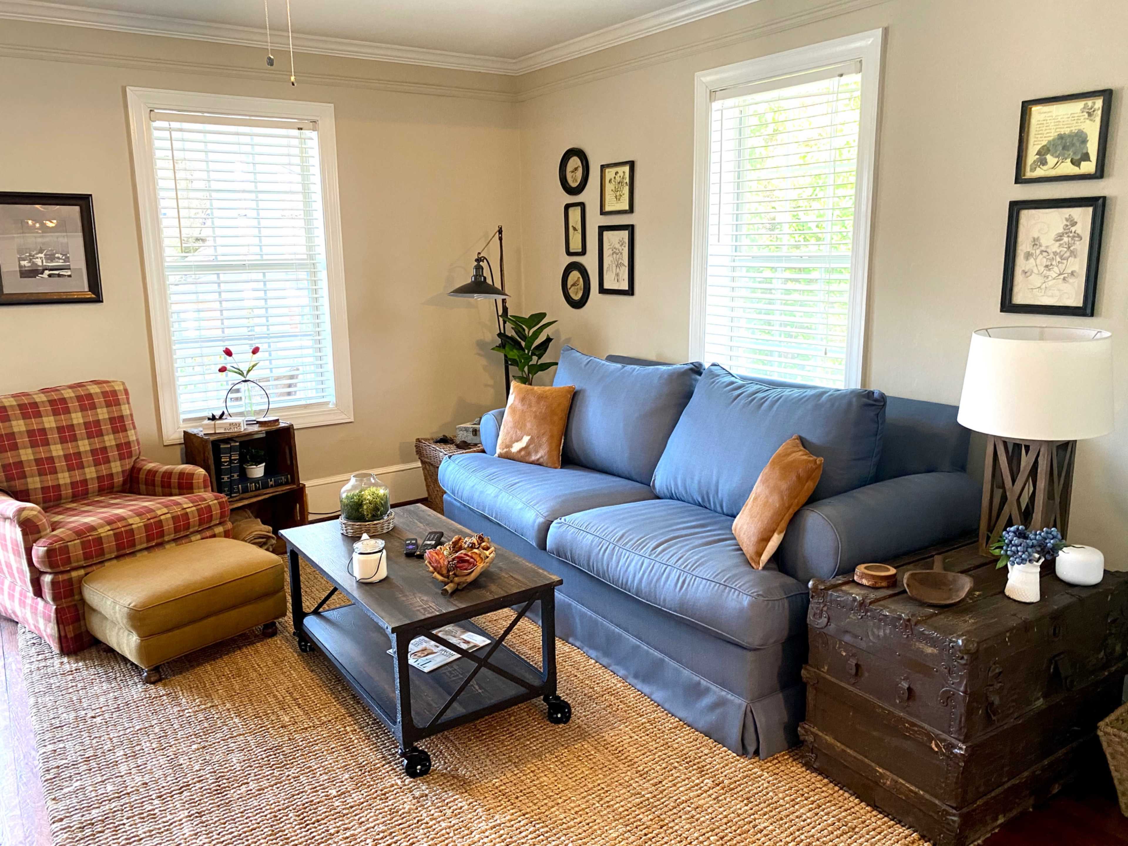 A cozy living room features a blue sofa, a plaid armchair, a coffee table on a jute rug, and various framed pictures on the walls.