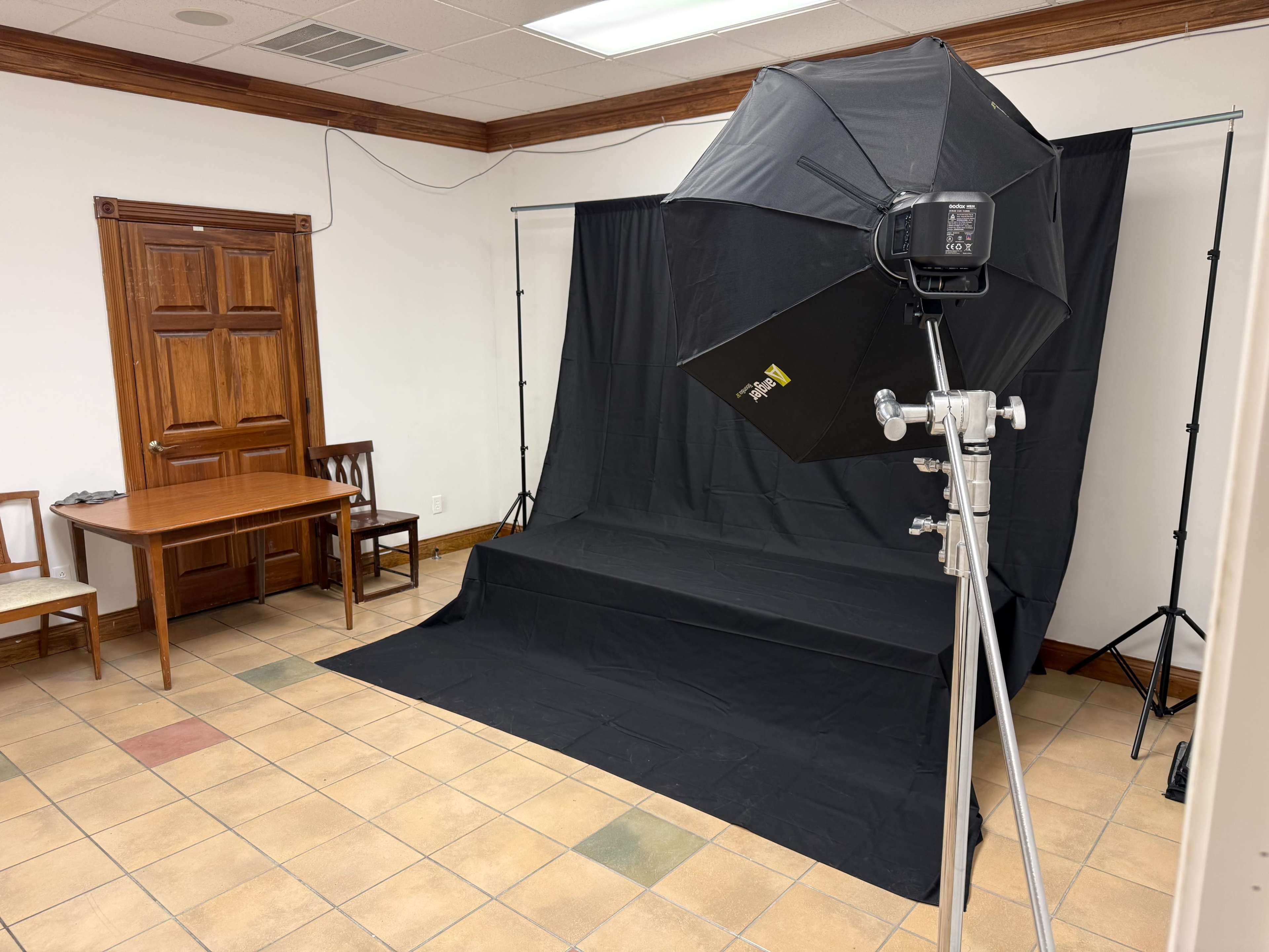 The image shows a simple photography studio setup featuring a black backdrop, a light stand with an umbrella light, a small table, and two chairs in a corner of the room.