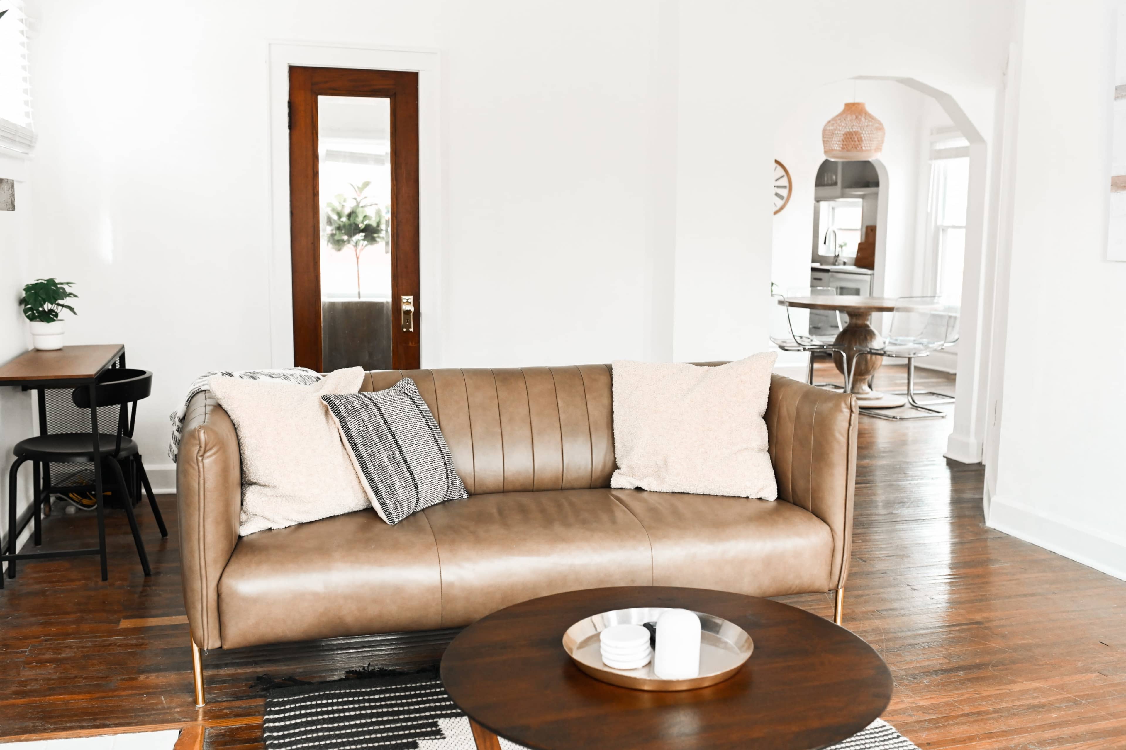 A leather sofa with two pillows sits in a bright living room, facing a round wooden coffee table.