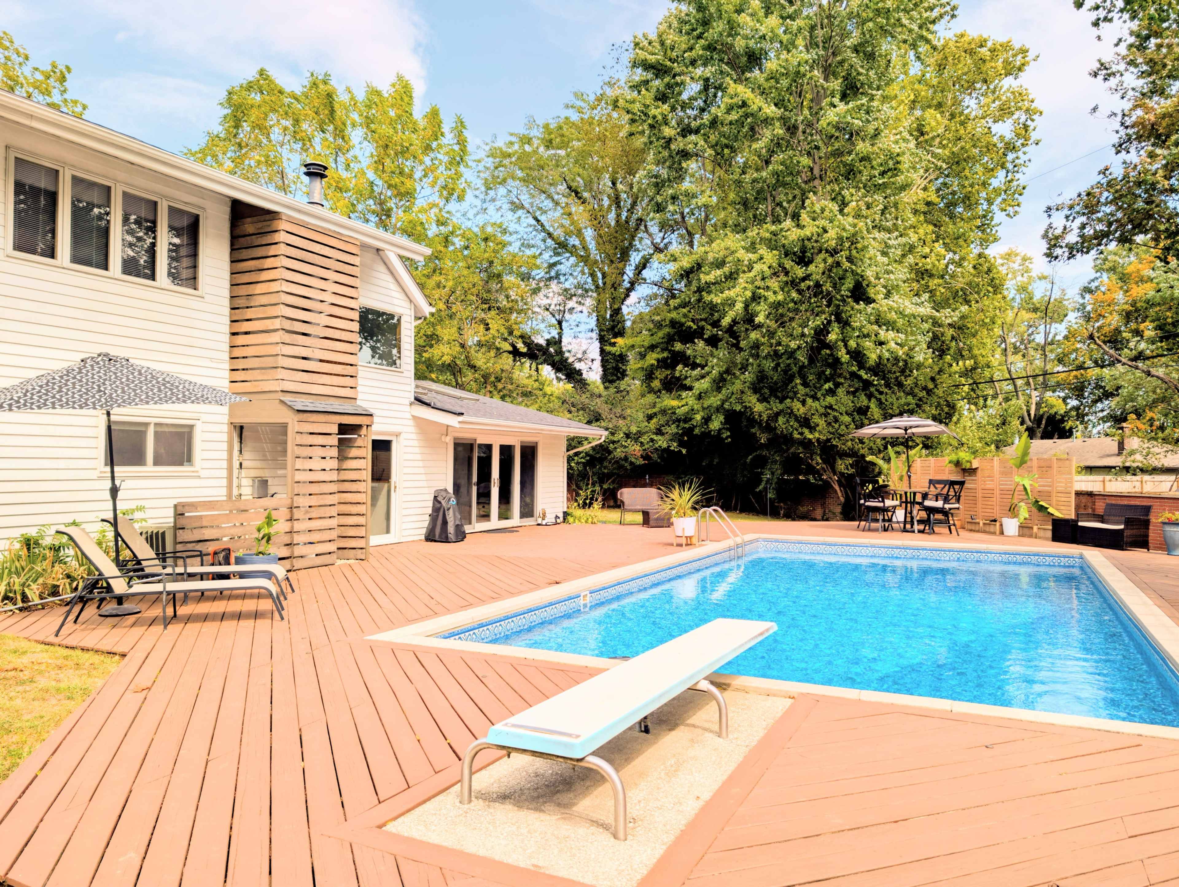 The image shows a house with a wooden deck surrounding a swimming pool, featuring lounge chairs and shade umbrellas.
