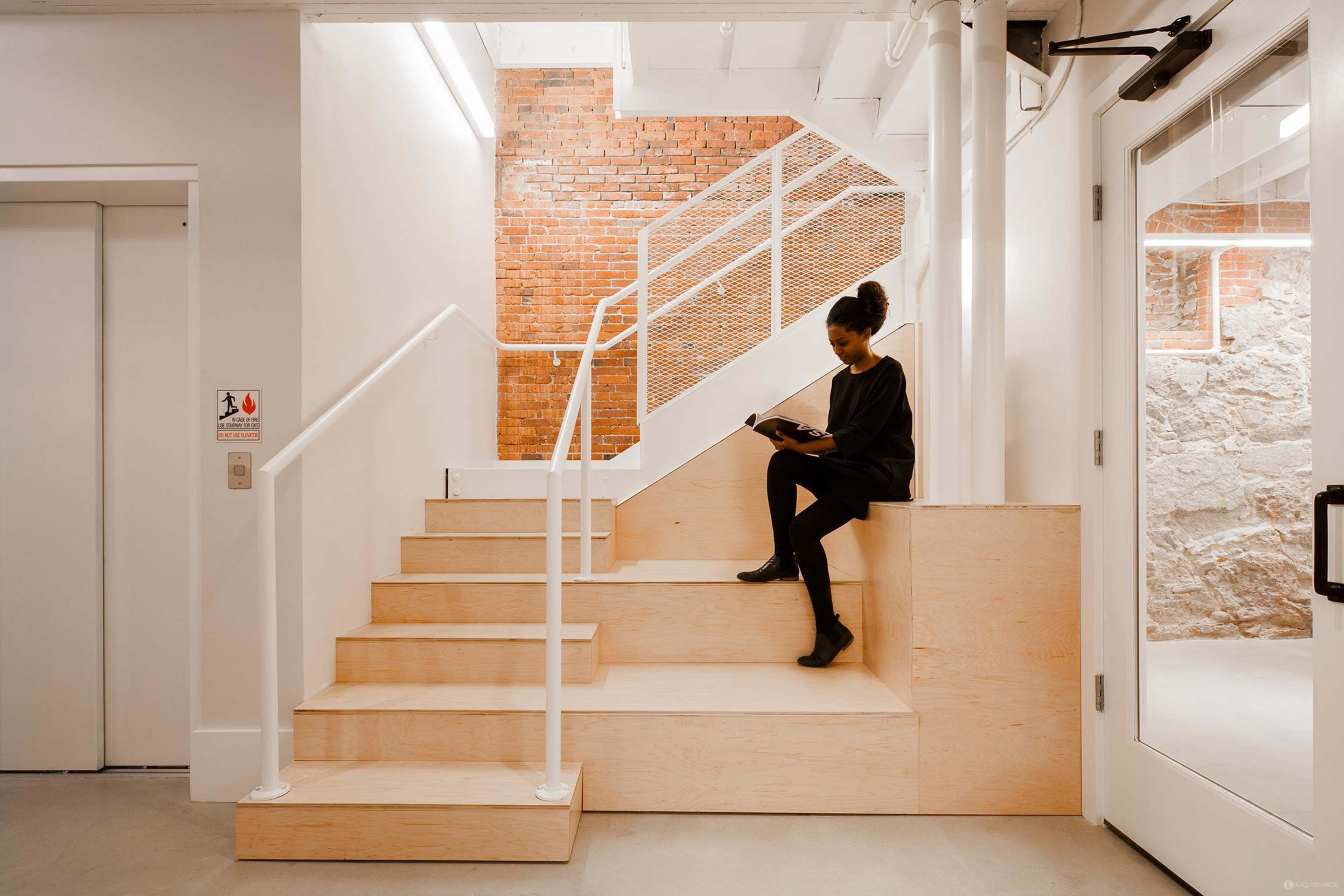 A woman sits on wooden steps in a modern building, reading a book while facing a stairway with exposed brick walls.