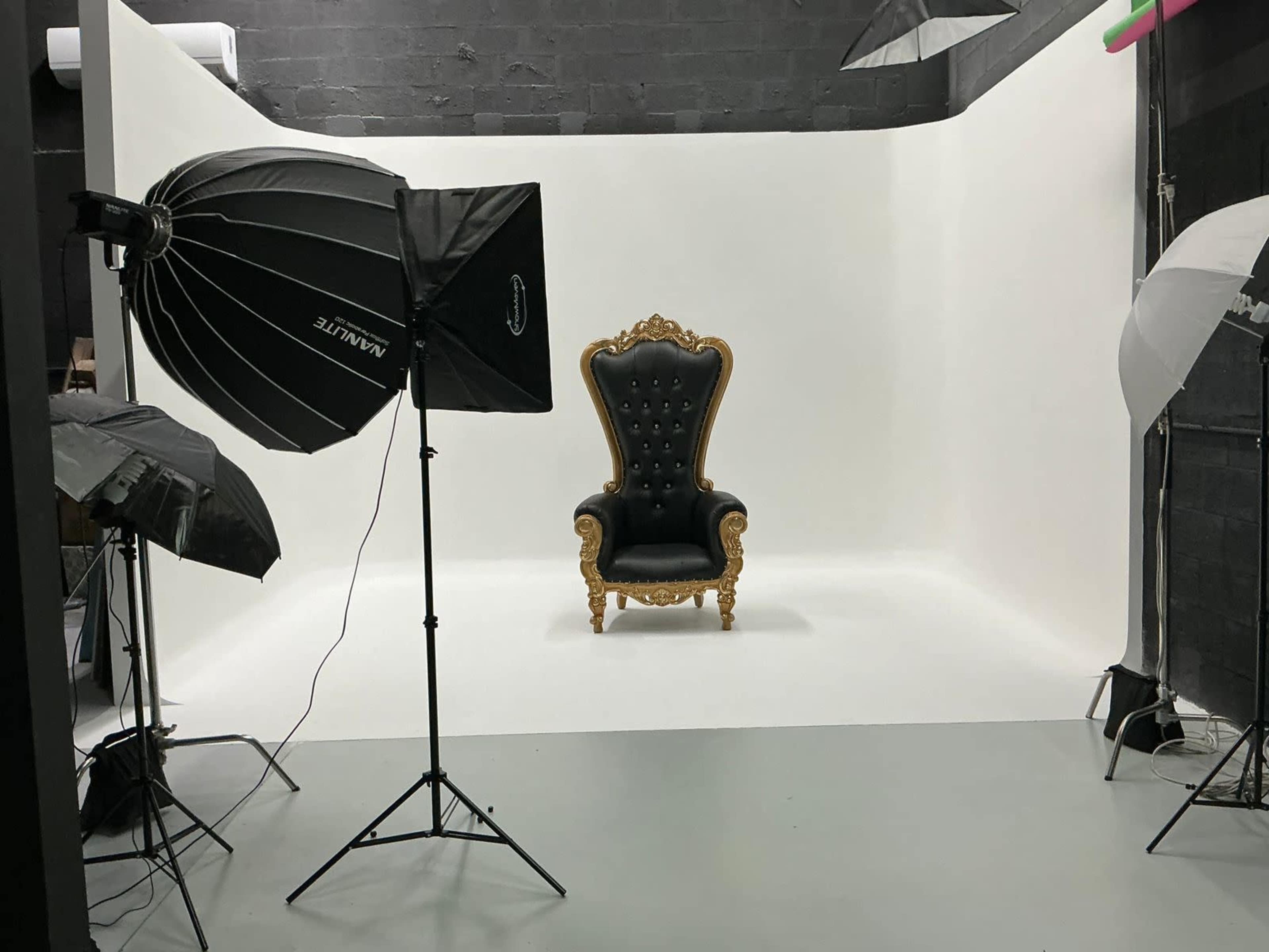 The image shows a photography studio with a large black and gold throne in front of a white backdrop, surrounded by studio lighting equipment.